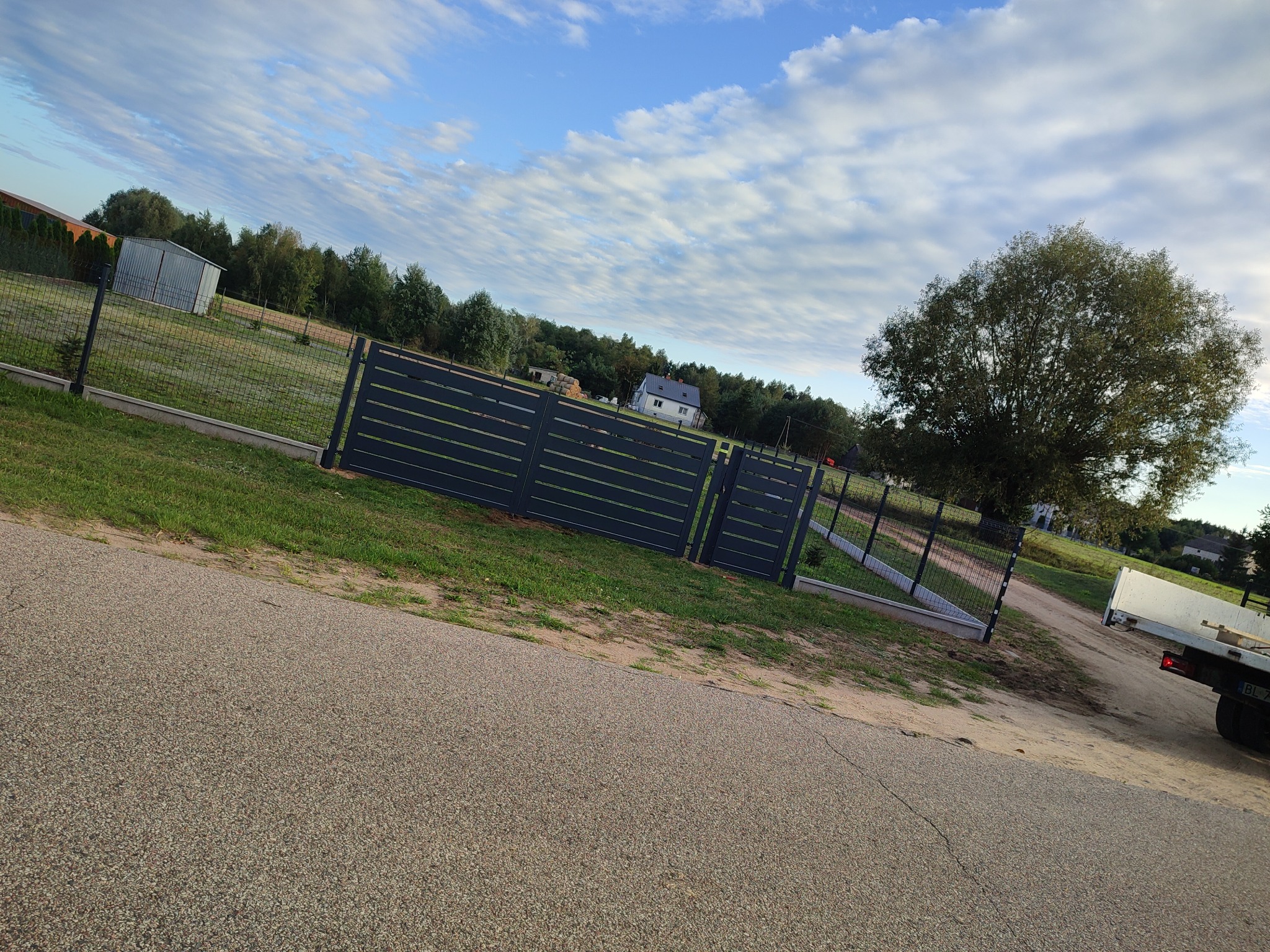 Nowoczesne, poziome ogrodzenie panelowe w kolorze grafitowym, z bramą wjazdową, na tle zielonego trawnika i błękitnego nieba z chmurami.