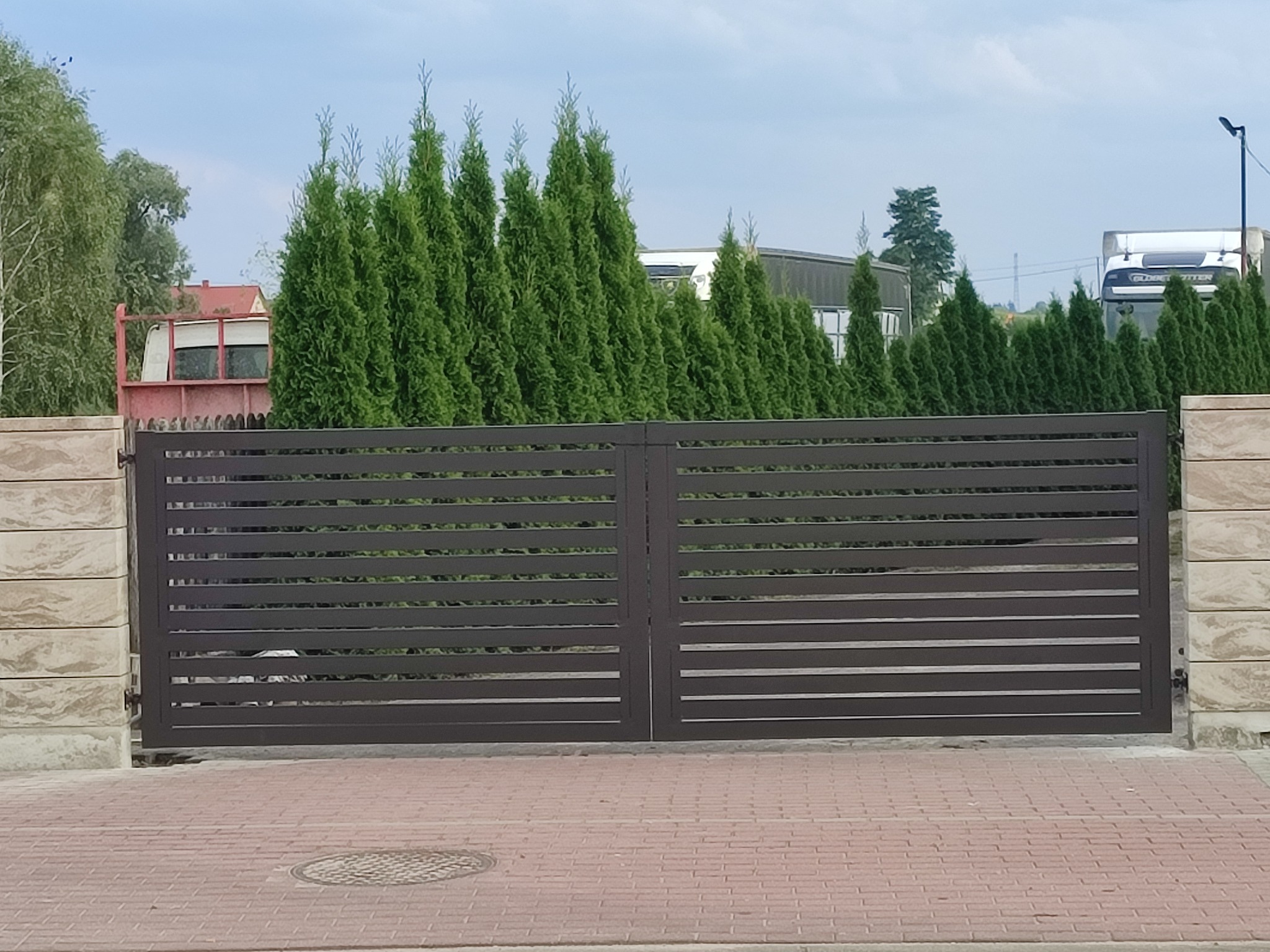 Nowoczesna, pozioma brama wjazdowa z metalowymi przęsłami, osadzona na kamiennych słupkach. W tle rząd wysokich, zielonych tuj i zaparkowany samochód ciężarowy.