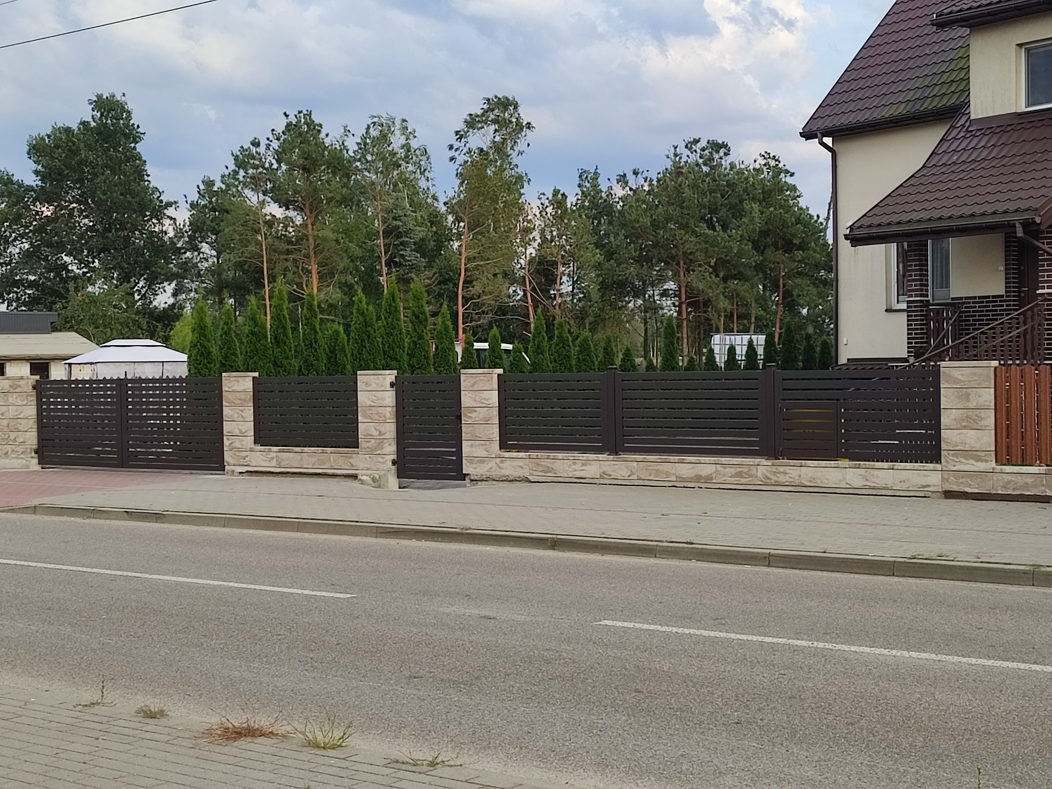 Nowoczesne ogrodzenie posesji z brązowymi panelami i kamiennymi słupkami, widok z ulicy. Brama wjazdowa i furtka w linii ogrodzenia. Tło stanowią drzewa i budynek.