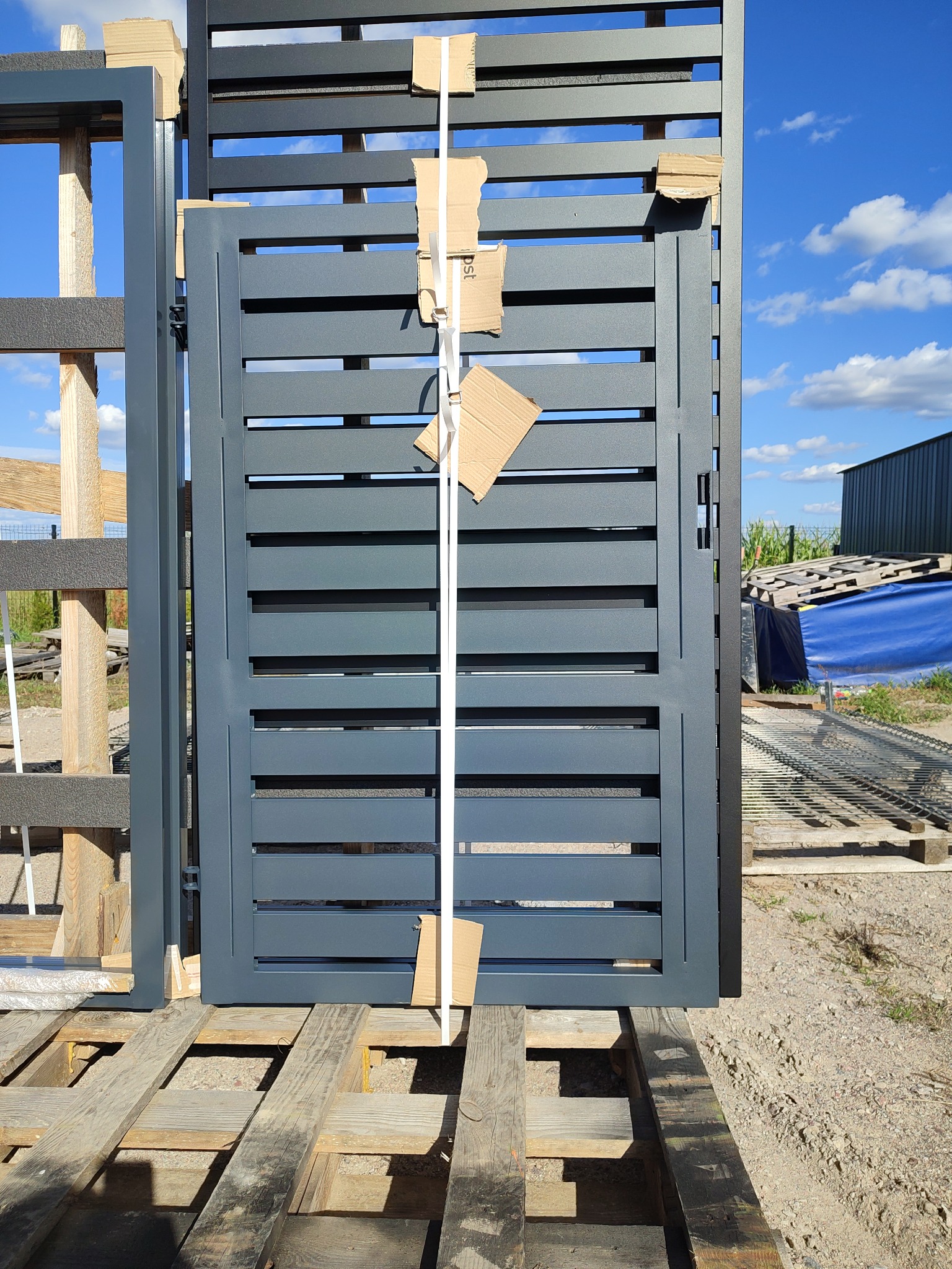 Szara, pozioma brama lamelowa na drewnianej palecie, zabezpieczona kartonem i taśmą. W tle błękitne niebo i budynek gospodarczy.
