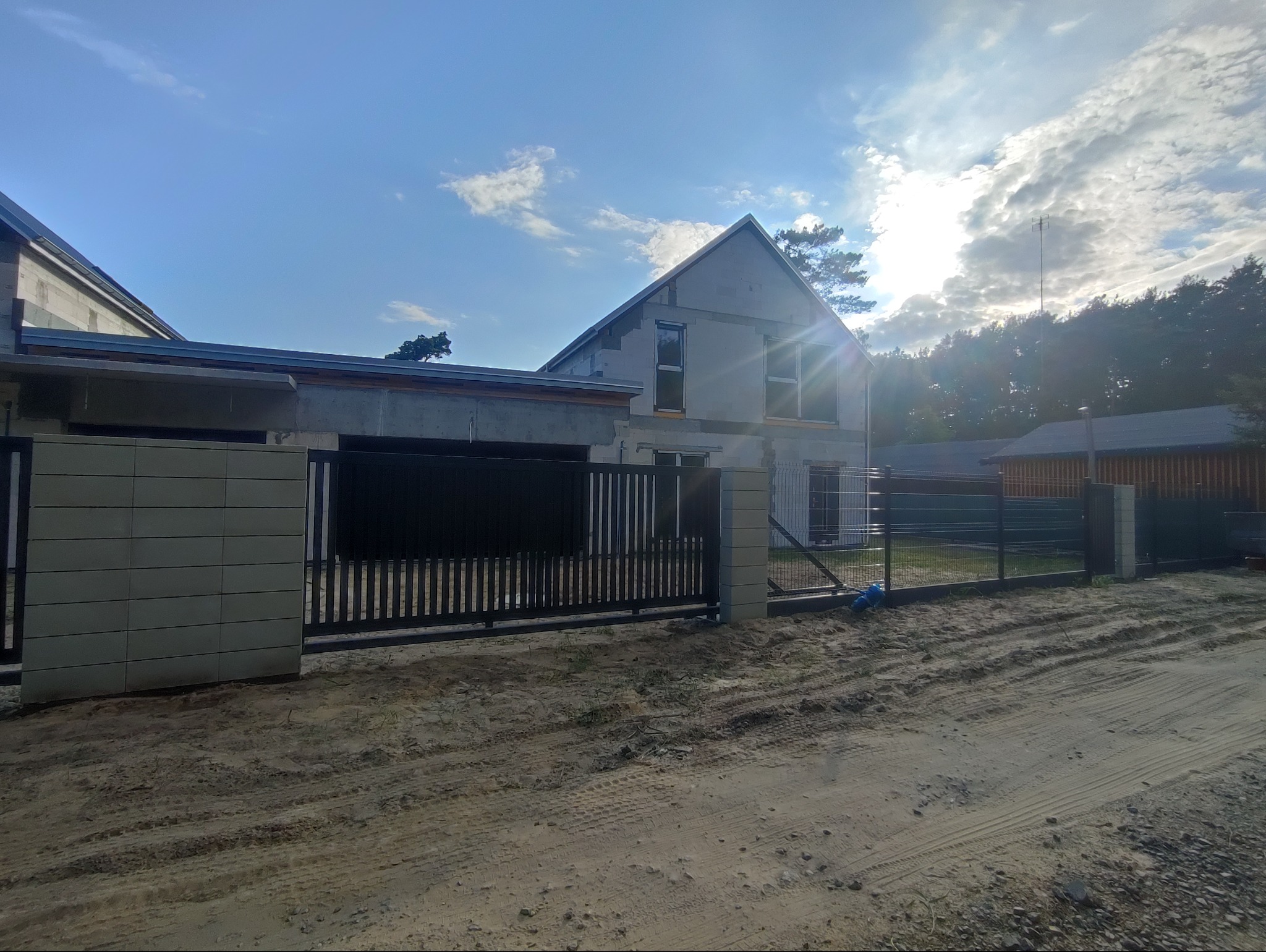 Nowoczesna, czarna brama przesuwna z pionowymi szczebelkami, zamontowana przy domu w trakcie budowy. Widoczne bloczki betonowe i fragment ogrodzenia panelowego.