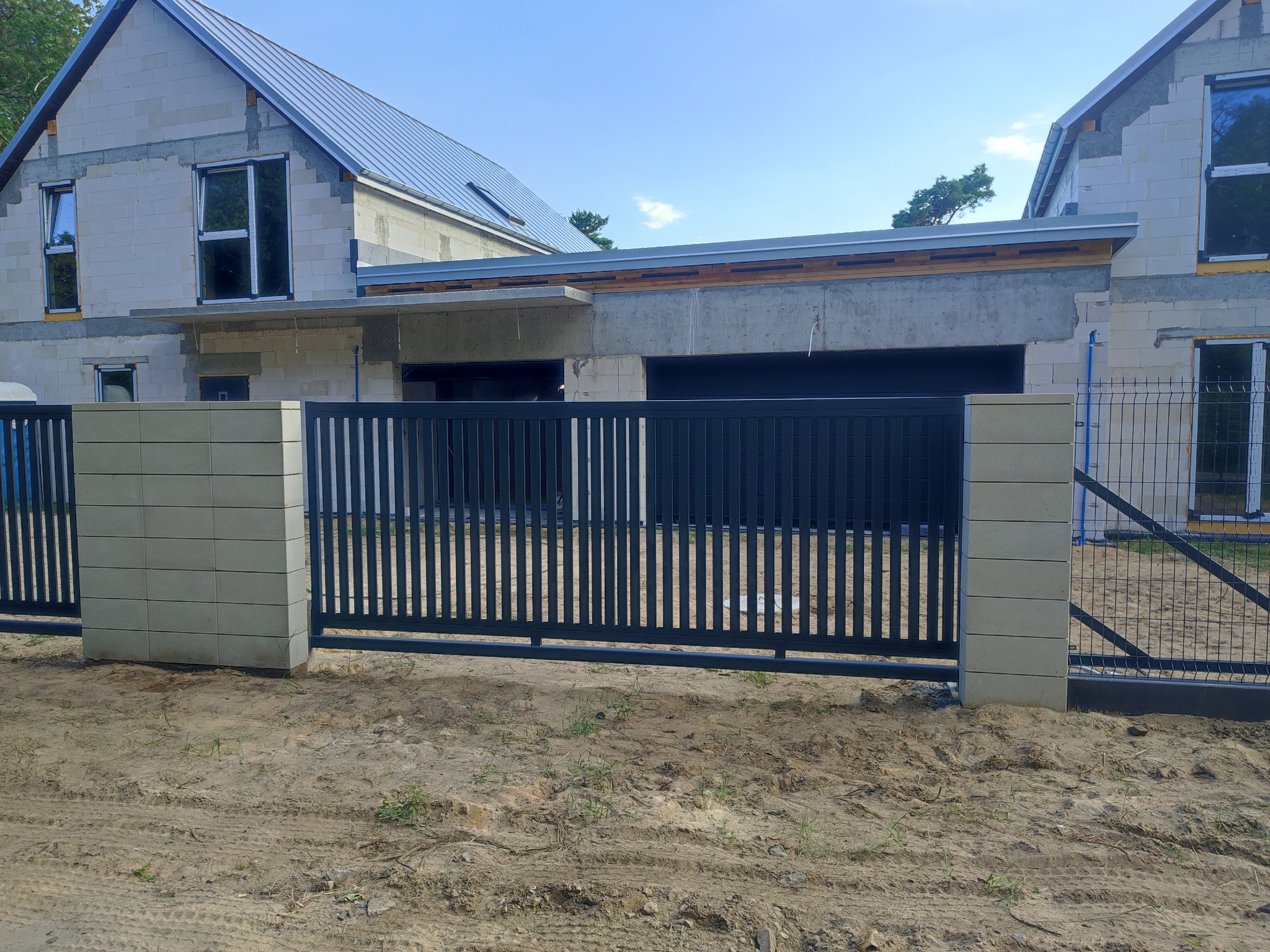 Nowoczesna, przesuwna brama w kolorze antracytu z betonowymi słupkami na tle domu w trakcie budowy. Widoczny fragment ogrodzenia panelowego.