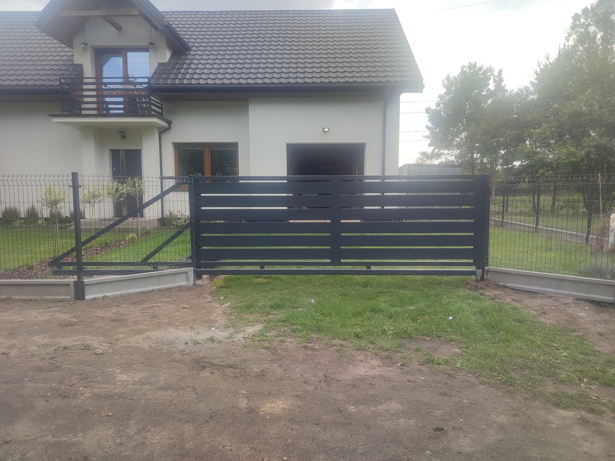 Nowoczesna, przesuwna brama wjazdowa z poziomych, grafitowych paneli na tle domu z ciemnym dachem i zieleni. Widoczne ogrodzenie panelowe.