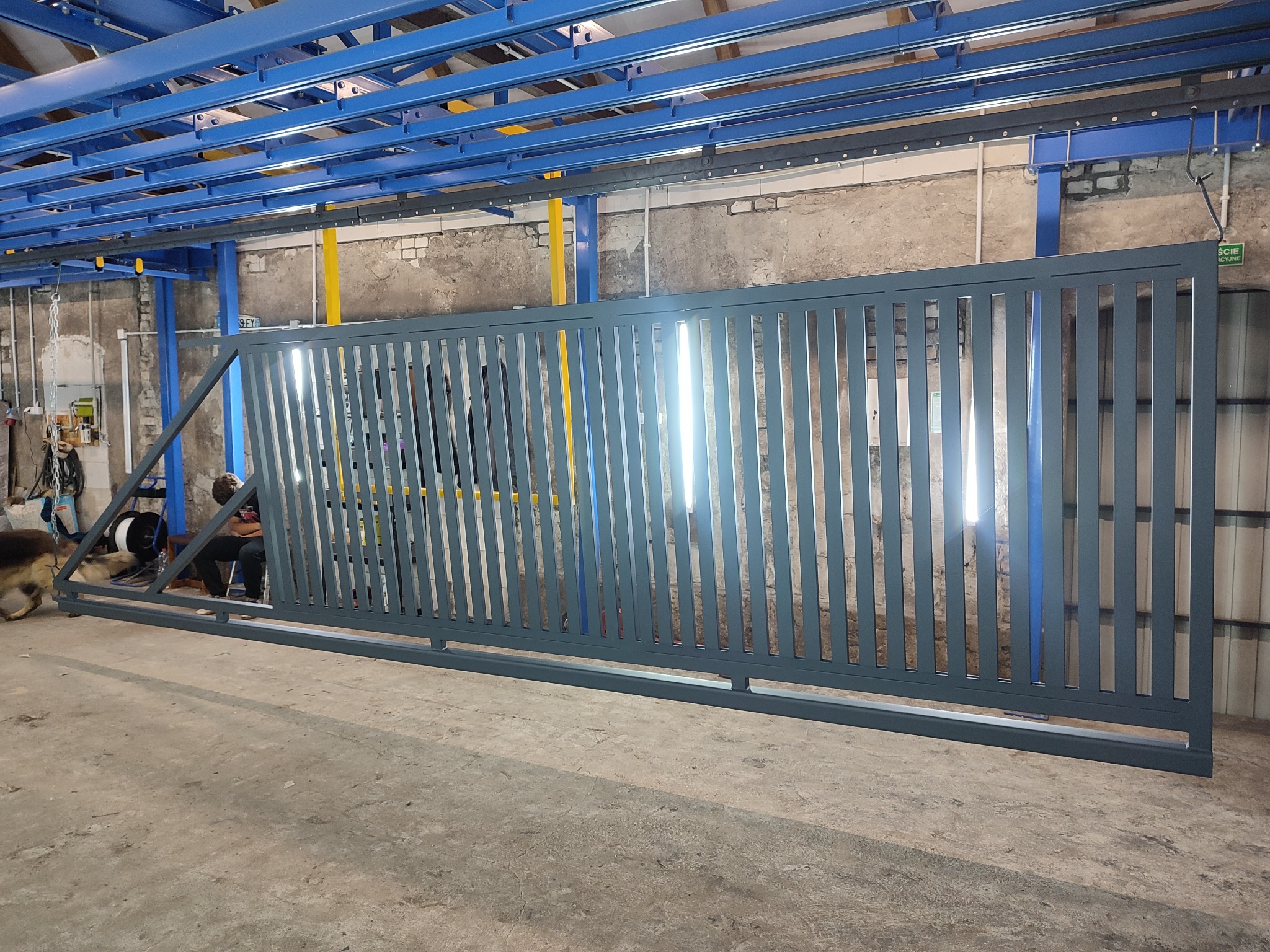 Szara, przesuwna brama metalowa z pionowymi szczebelkami, widoczna w hali produkcyjnej z niebieską konstrukcją sufitową i fragmentem osoby w tle.