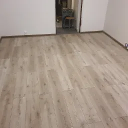 Ułożone panele podłogowe w jasnym odcieniu drewna, widoczne białe ściany i fragment przejścia do innego pomieszczenia z widocznym głośnikiem i krzesłem.