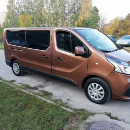 Brązowy van Renault Trafic z przyciemnianymi szybami, zaparkowany na betonowym placu z zielenią, w tle inne samochody i budynek.