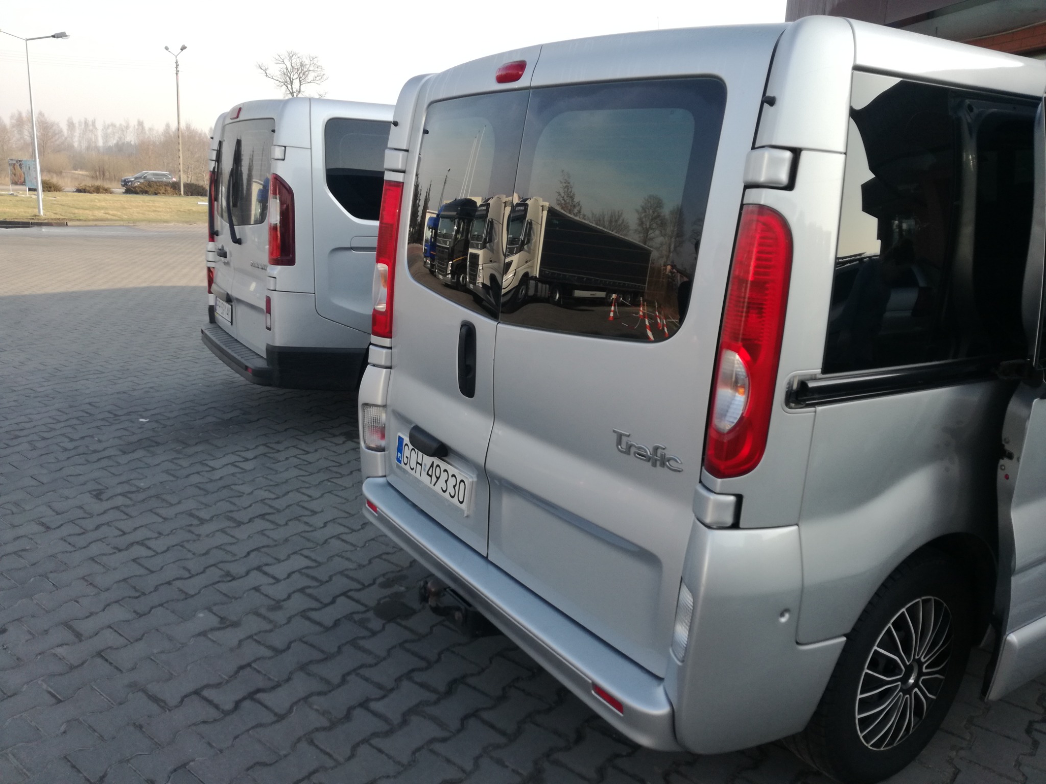 Dwa srebrne busy Renault Trafic zaparkowane na brukowanym placu; w szybie jednego z nich odbijają się zaparkowane ciężarówki, a drzwi boczne są otwarte.