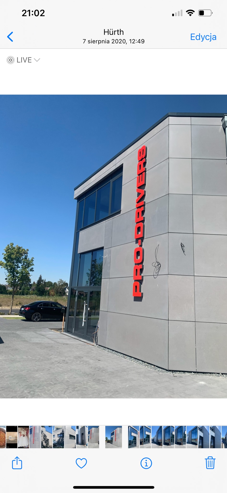 Budynek biurowy z szarą elewacją z paneli i czerwonym pionowym napisem 'PRO-DRIVERS', widoczne zarysowania na elewacji, zaparkowany czarny samochód osobowy, błękitne niebo.