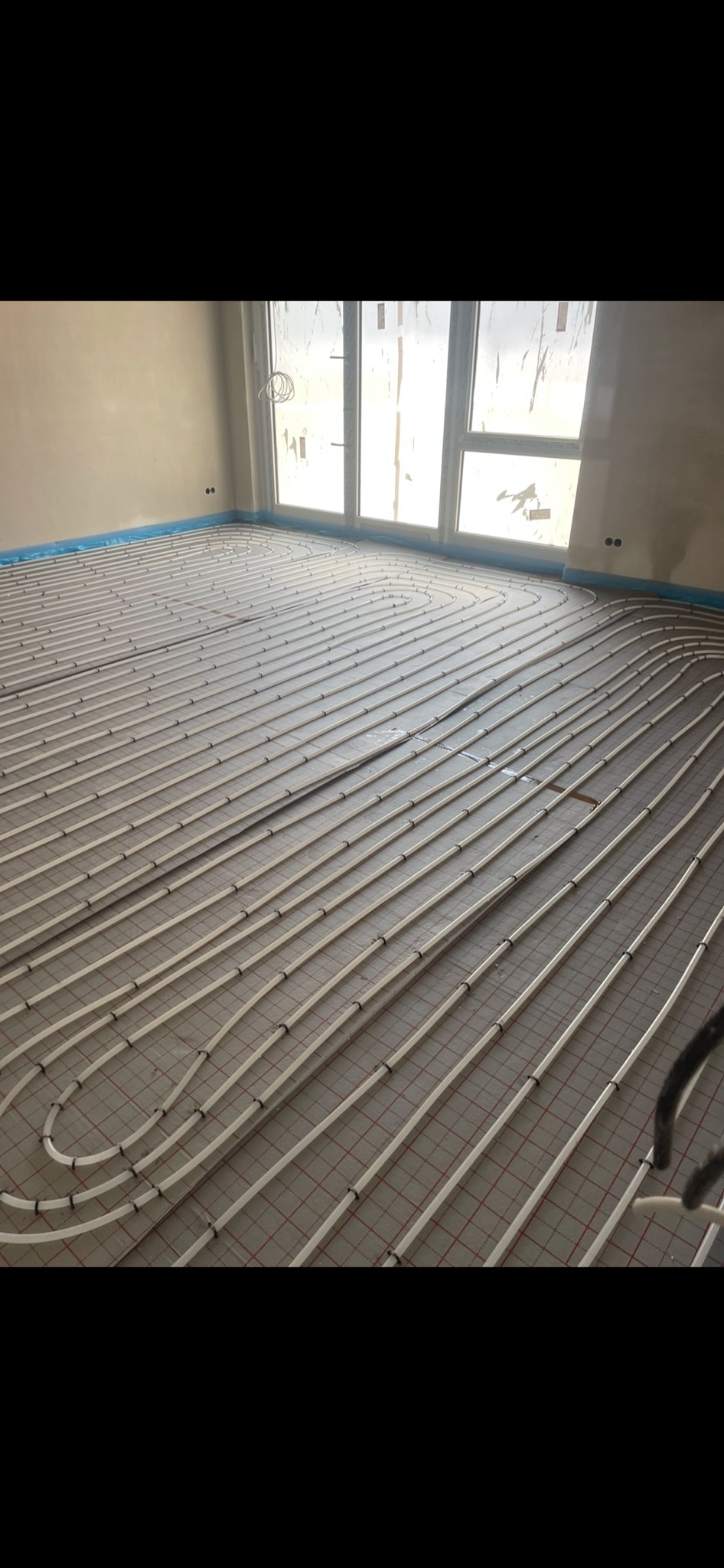 Rozłożone rury ogrzewania podłogowego na siatce w surowym wnętrzu z dużym oknem, przygotowane do zalania wylewką.