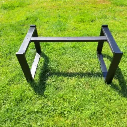 Czarna, metalowa podstawa stołu na trawie w słoneczny dzień. Konstrukcja spawana z profili zamkniętych.