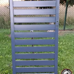 Szara, metalowa brama wjazdowa z poziomymi panelami, oparta o drewniane deski na trawniku, widoczna przez szczeliny siatka ogrodzeniowa i zieleń, w prawym dolnym rogu logo firmy.