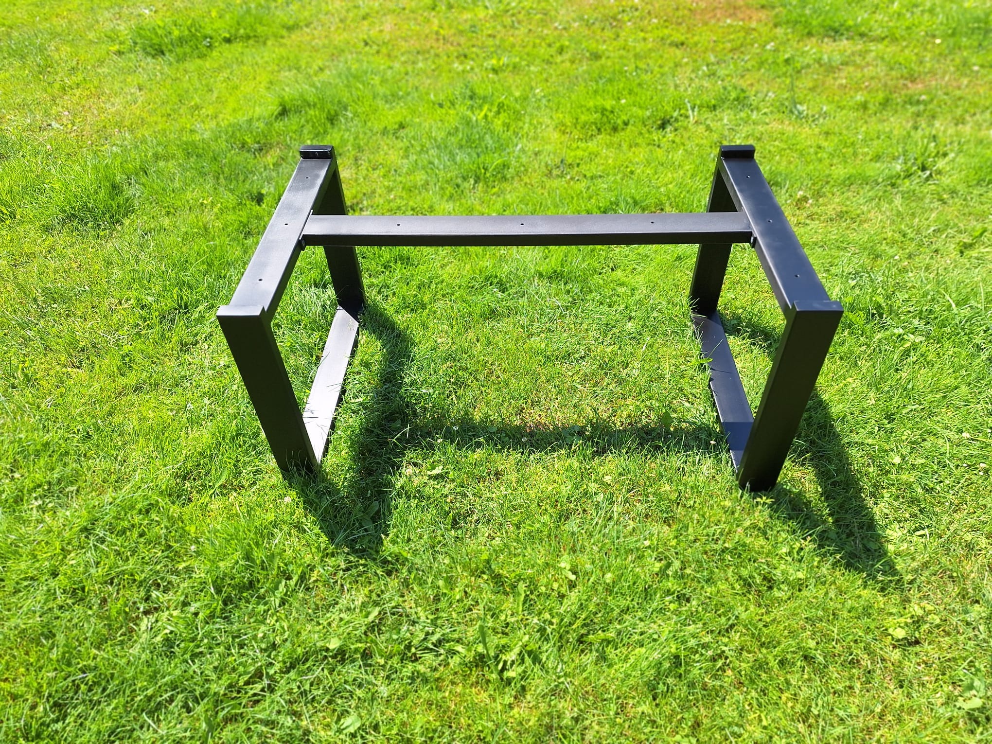 Czarna, metalowa podstawa stołu na trawie w słoneczny dzień. Konstrukcja spawana z profili zamkniętych.