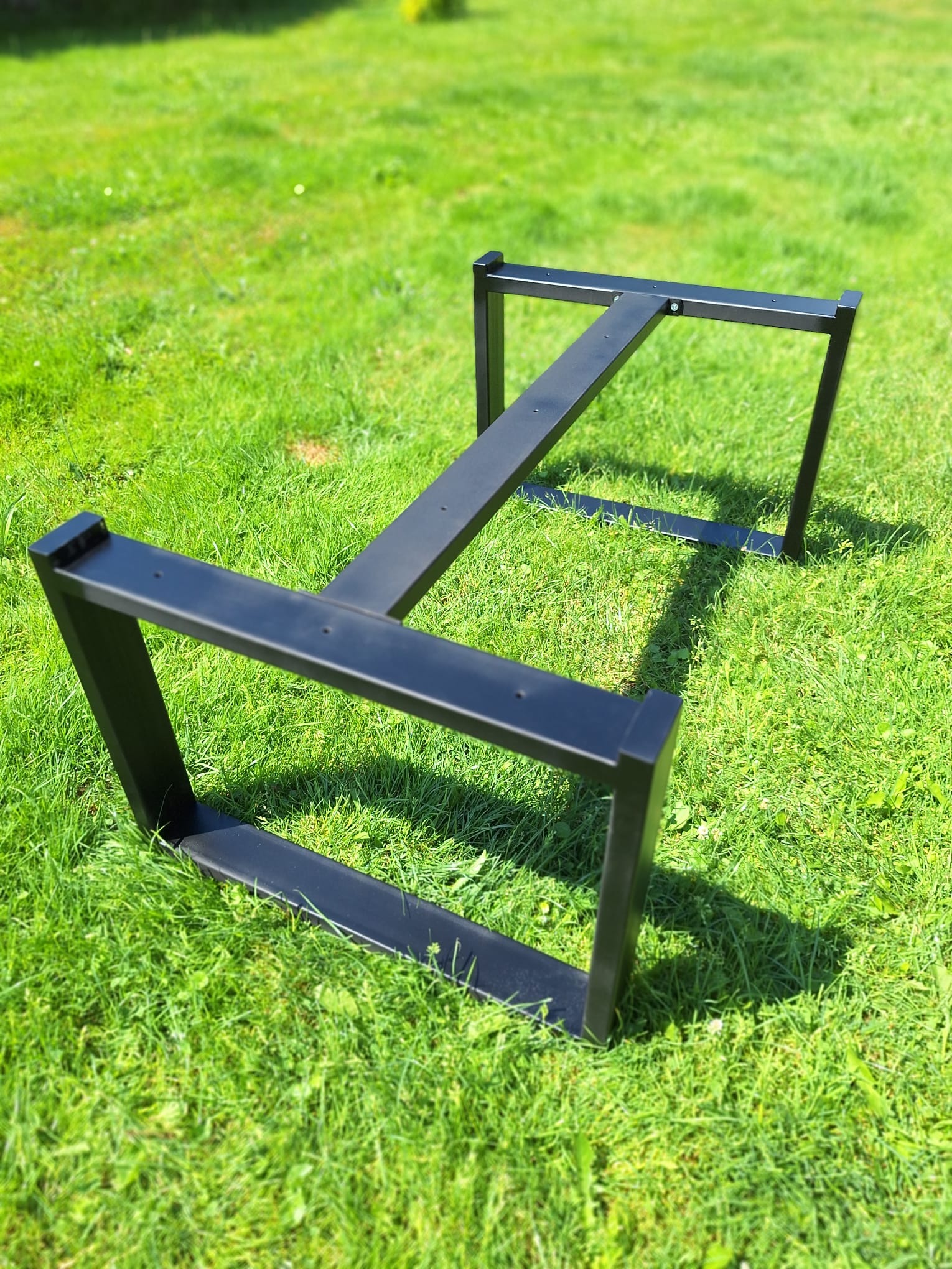 Metalowa podstawa stołu o prostym, geometrycznym kształcie, pomalowana na czarno, leży na zielonej trawie w słoneczny dzień.