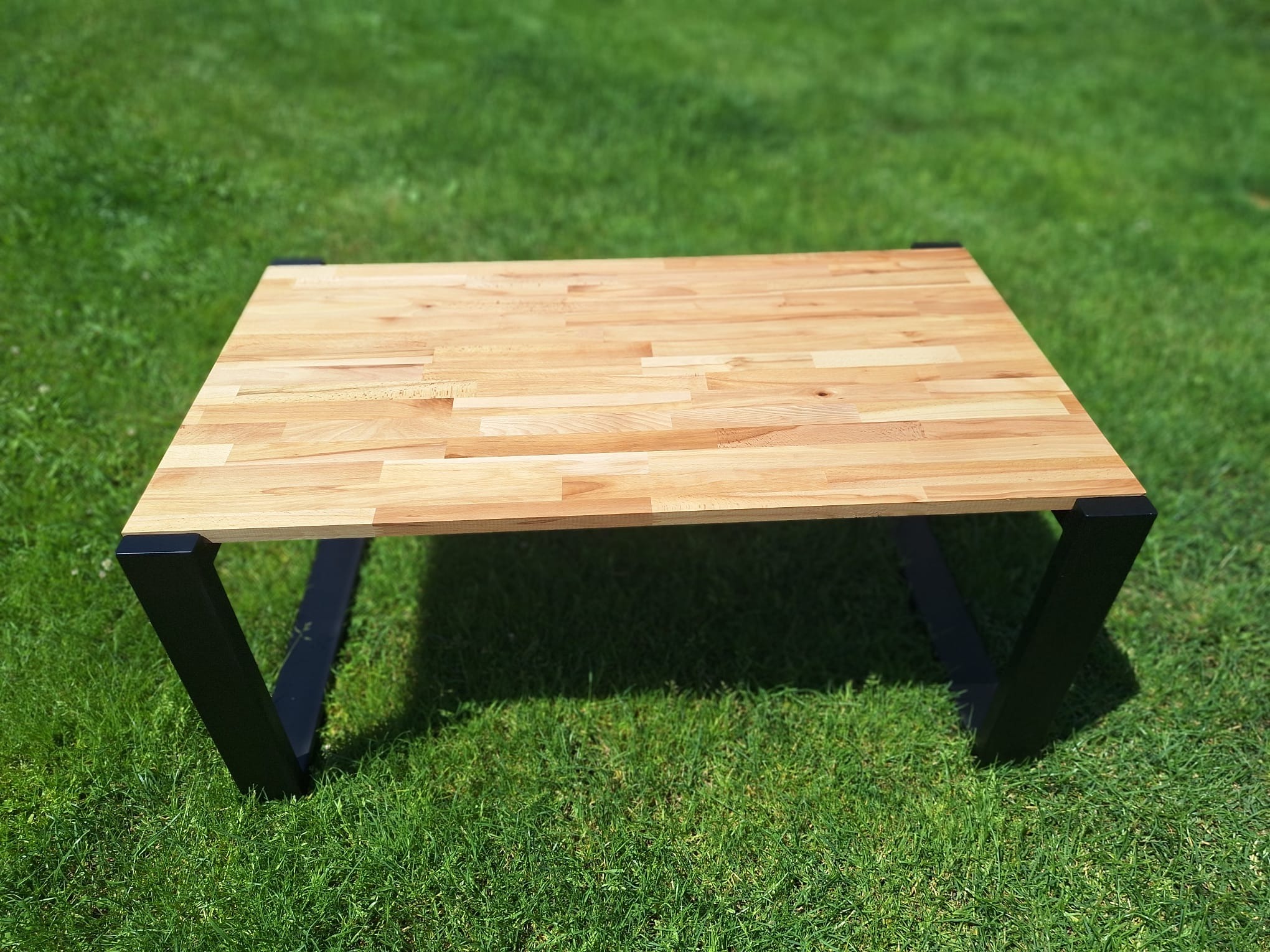 Stolik kawowy z blatem z jasnego drewna klejonego i czarnymi, metalowymi nogami w kształcie litery V, ustawiony na zielonej trawie w słoneczny dzień.