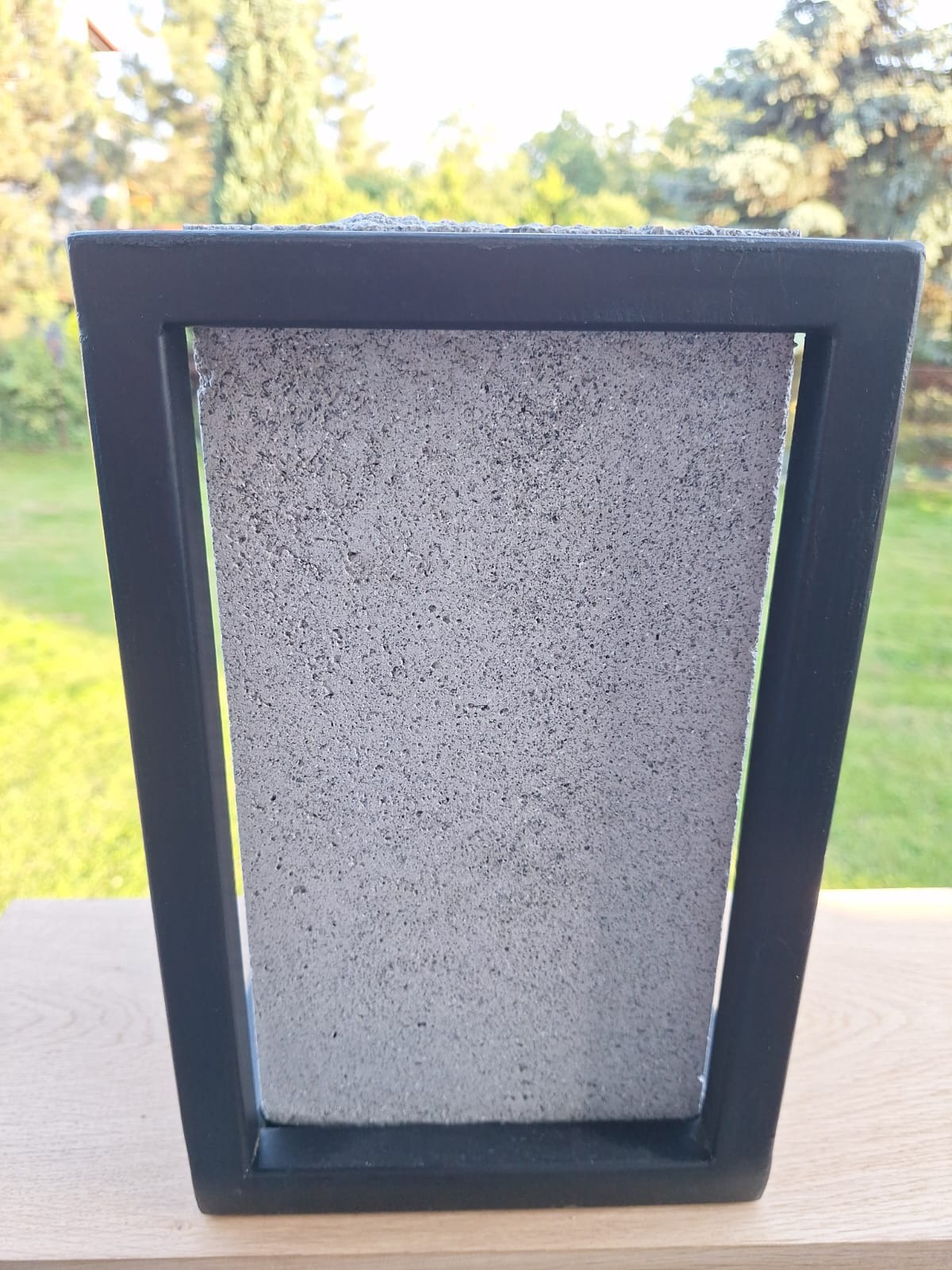 Nowoczesna donica betonowa z czarną, metalową ramą, ustawiona na drewnianym blacie na tle zieleni ogrodu.