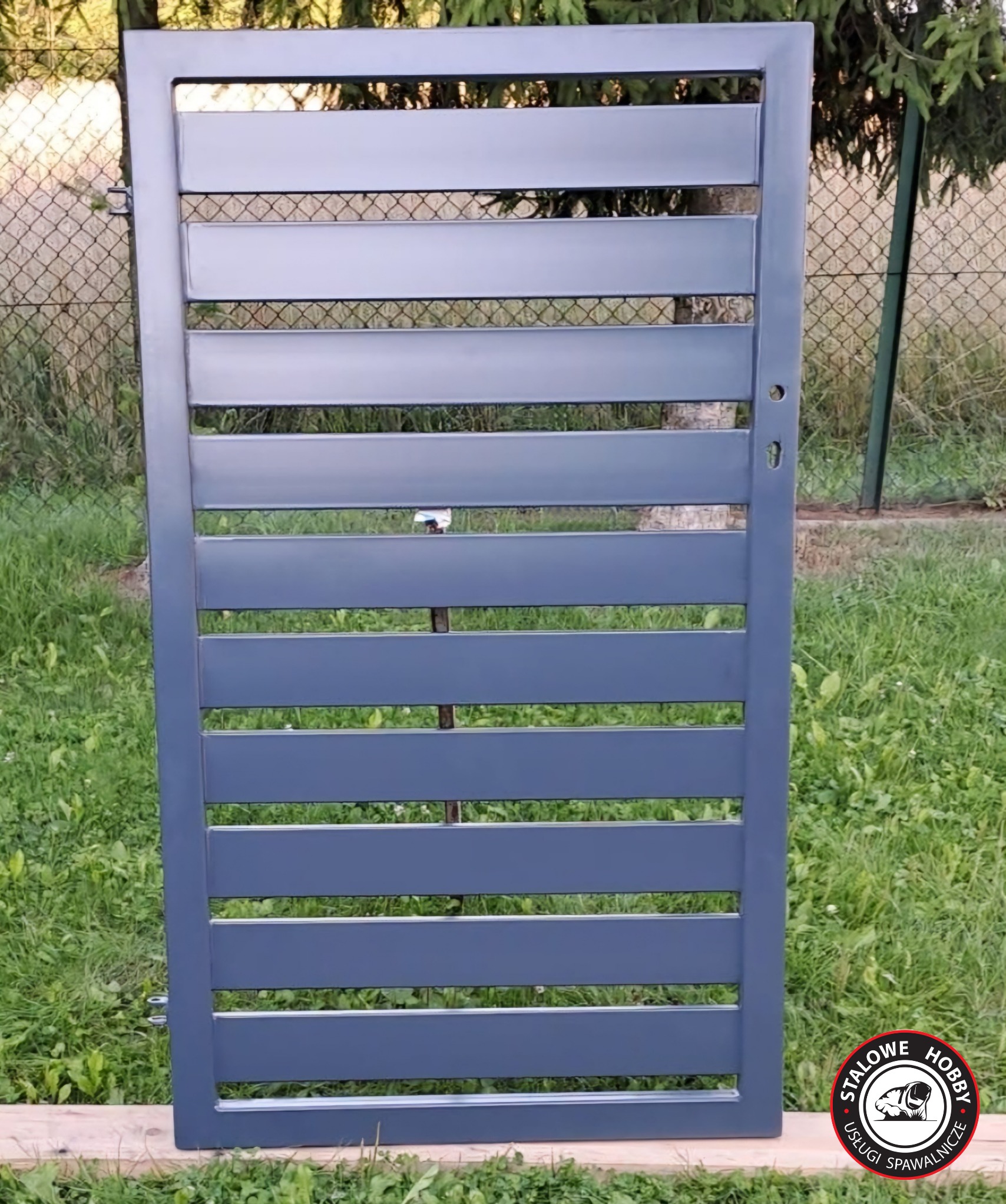 Szara, metalowa brama wjazdowa z poziomymi panelami, oparta o drewniane deski na trawniku, widoczna przez szczeliny siatka ogrodzeniowa i zieleń, w prawym dolnym rogu logo firmy.