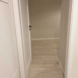 USŁUGI REMONTOWO-BUDOWLANE DAMIAN STĘPIEŃ - Otwarta biała rama drzwiowa z czarną klamką, widok na jasne panele podłogowe i beżową ścianę w tle.