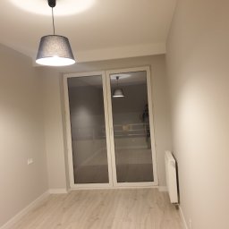 USŁUGI REMONTOWO-BUDOWLANE DAMIAN STĘPIEŃ - Pomalowane na jasny beż pokój z drewnianą podłogą, widoczne okno balkonowe z białymi ramami, grzejnik i lampa wisząca z szarym abażurem.
