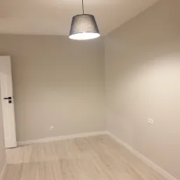 Pomalowane na beżowo puste pomieszczenie z panelami podłogowymi, białą listwą przypodłogową i szarą lampą wiszącą z materiałowym abażurem.