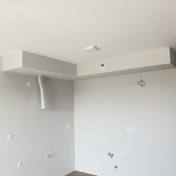 USŁUGI REMONTOWO-BUDOWLANE DAMIAN STĘPIEŃ - Surowe wnętrze w trakcie remontu: widoczne wystające kable, rury wentylacyjne i gniazdka elektryczne na świeżo pomalowanych, szarych ścianach z drewnianą podłogą.