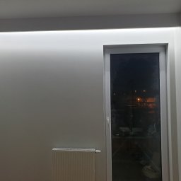 USŁUGI REMONTOWO-BUDOWLANE DAMIAN STĘPIEŃ - Białe, pomalowane na gładko ściany z oświetleniem LED podsufitowym, okno PCV z widokiem na nocne miasto, grzejnik panelowy pod oknem i gniazdko elektryczne na ścianie.