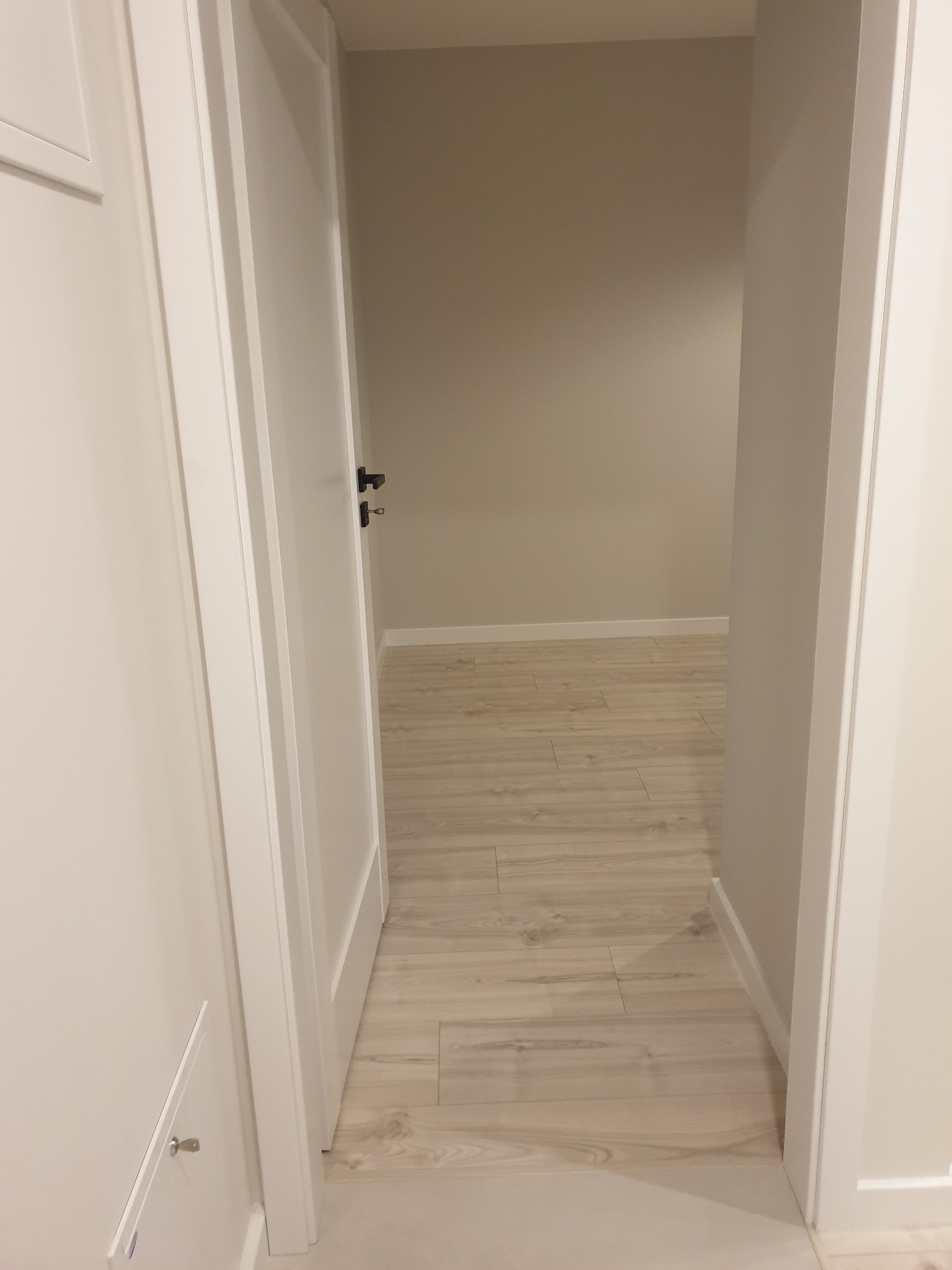 Otwarta biała rama drzwiowa z czarną klamką, widok na jasne panele podłogowe i beżową ścianę w tle.