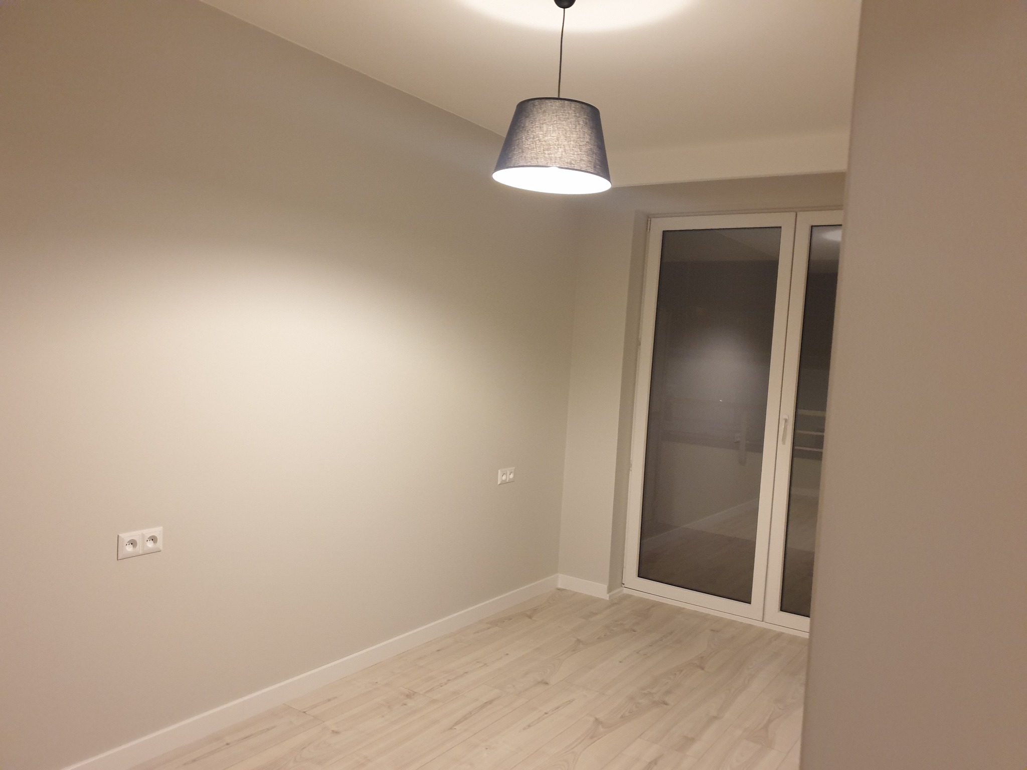 Pomalowane na jasny odcień szarości ściany w pustym pokoju z drewnopodobną podłogą, widoczne gniazdka elektryczne i lampa z abażurem.