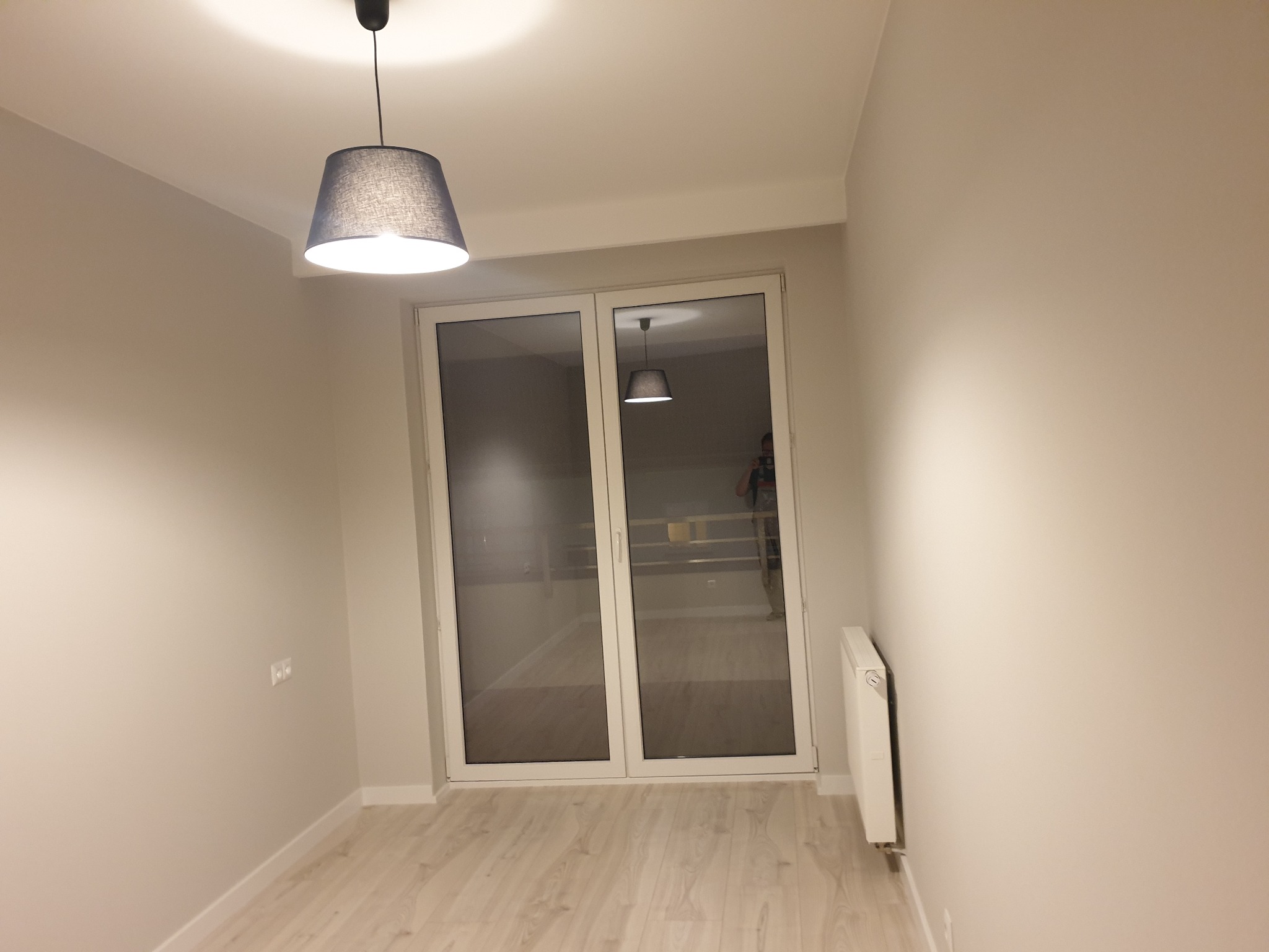 Pomalowane na jasny beż pokój z drewnianą podłogą, widoczne okno balkonowe z białymi ramami, grzejnik i lampa wisząca z szarym abażurem.