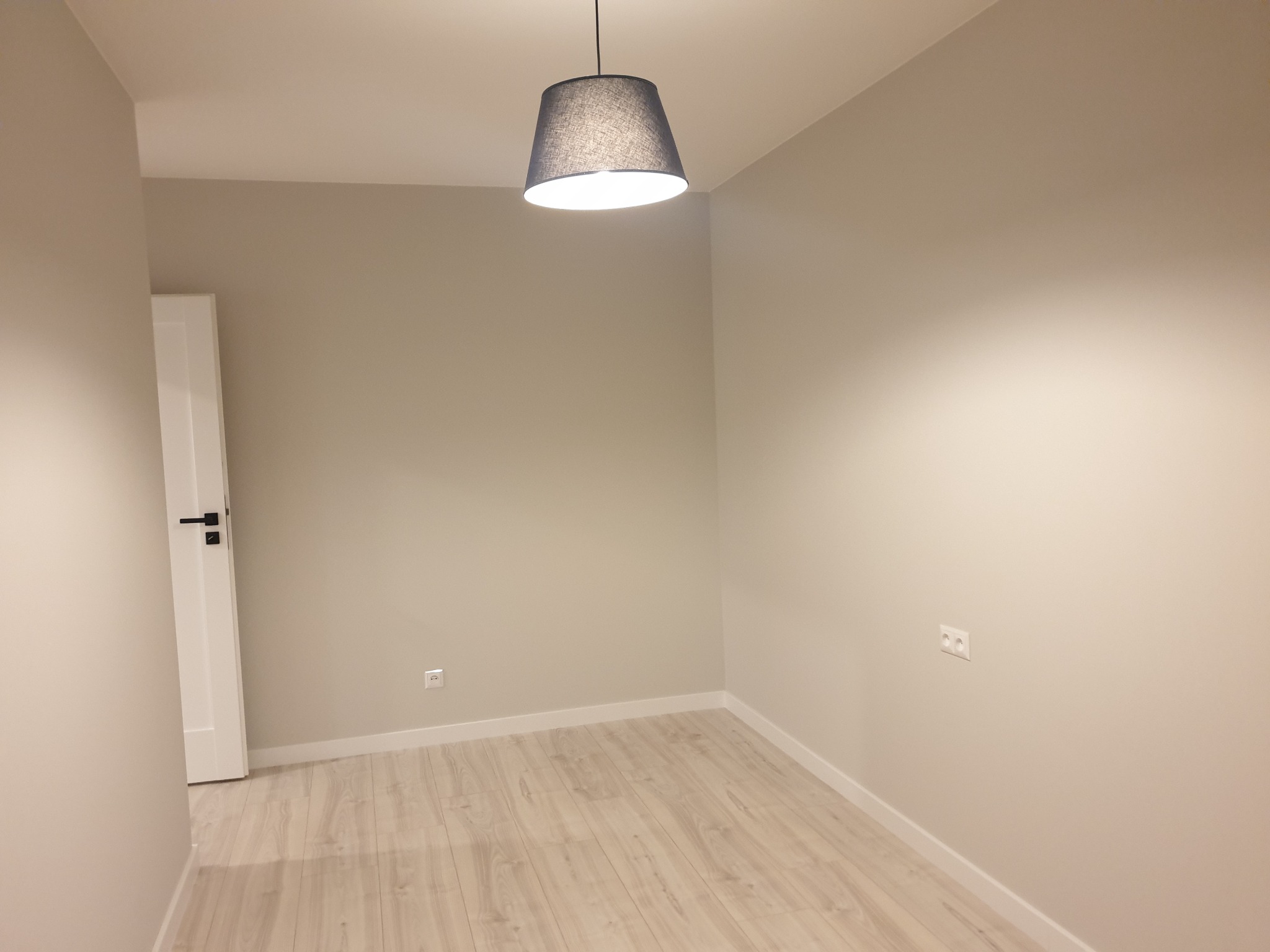 Pomalowane na beżowo puste pomieszczenie z panelami podłogowymi, białą listwą przypodłogową i szarą lampą wiszącą z materiałowym abażurem.