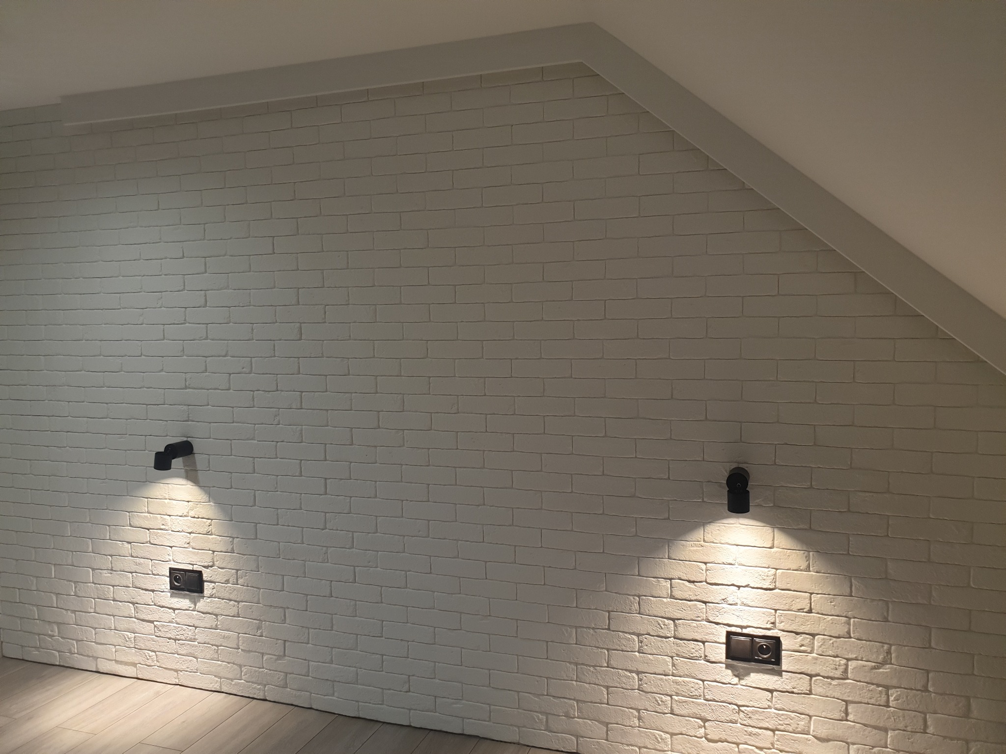 Ściana z białej cegły w pokoju na poddaszu, oświetlona dwoma czarnymi lampami ściennymi, z widocznymi gniazdkami elektrycznymi.