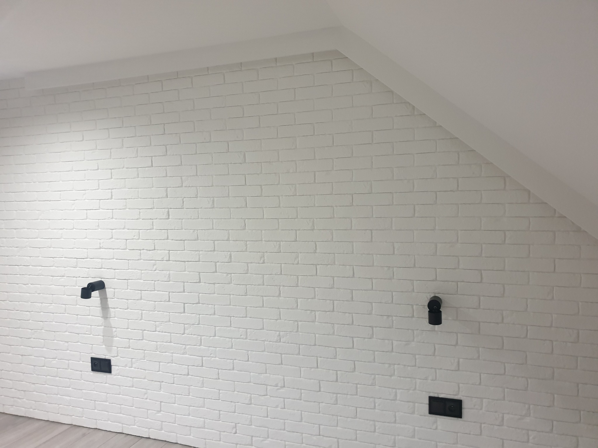 Ściana w pokoju na poddaszu wykończona białymi cegłami, z widocznymi czarnymi elementami instalacji elektrycznej i oświetleniowej.
