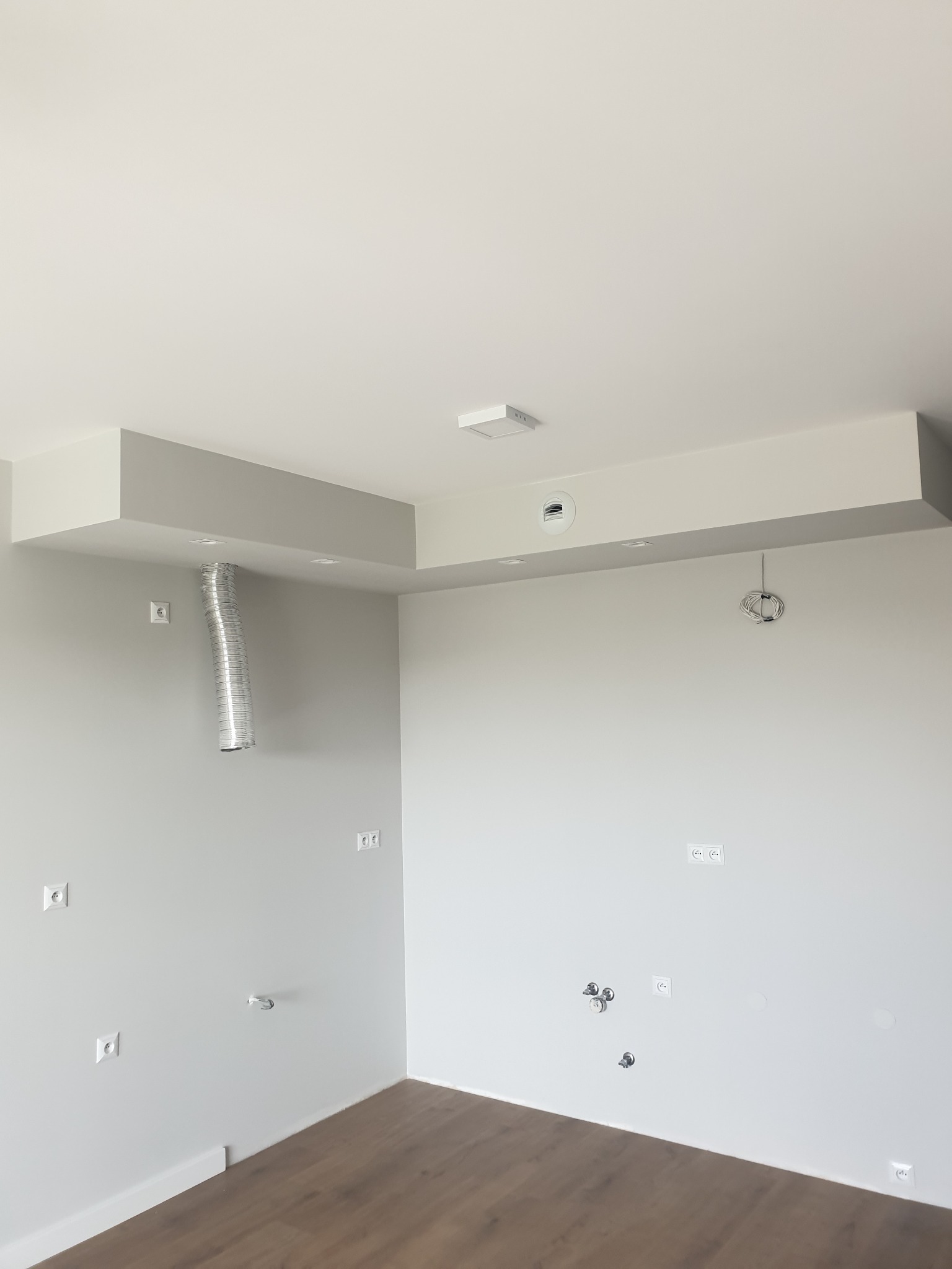 Surowe wnętrze w trakcie remontu: widoczne wystające kable, rury wentylacyjne i gniazdka elektryczne na świeżo pomalowanych, szarych ścianach z drewnianą podłogą.