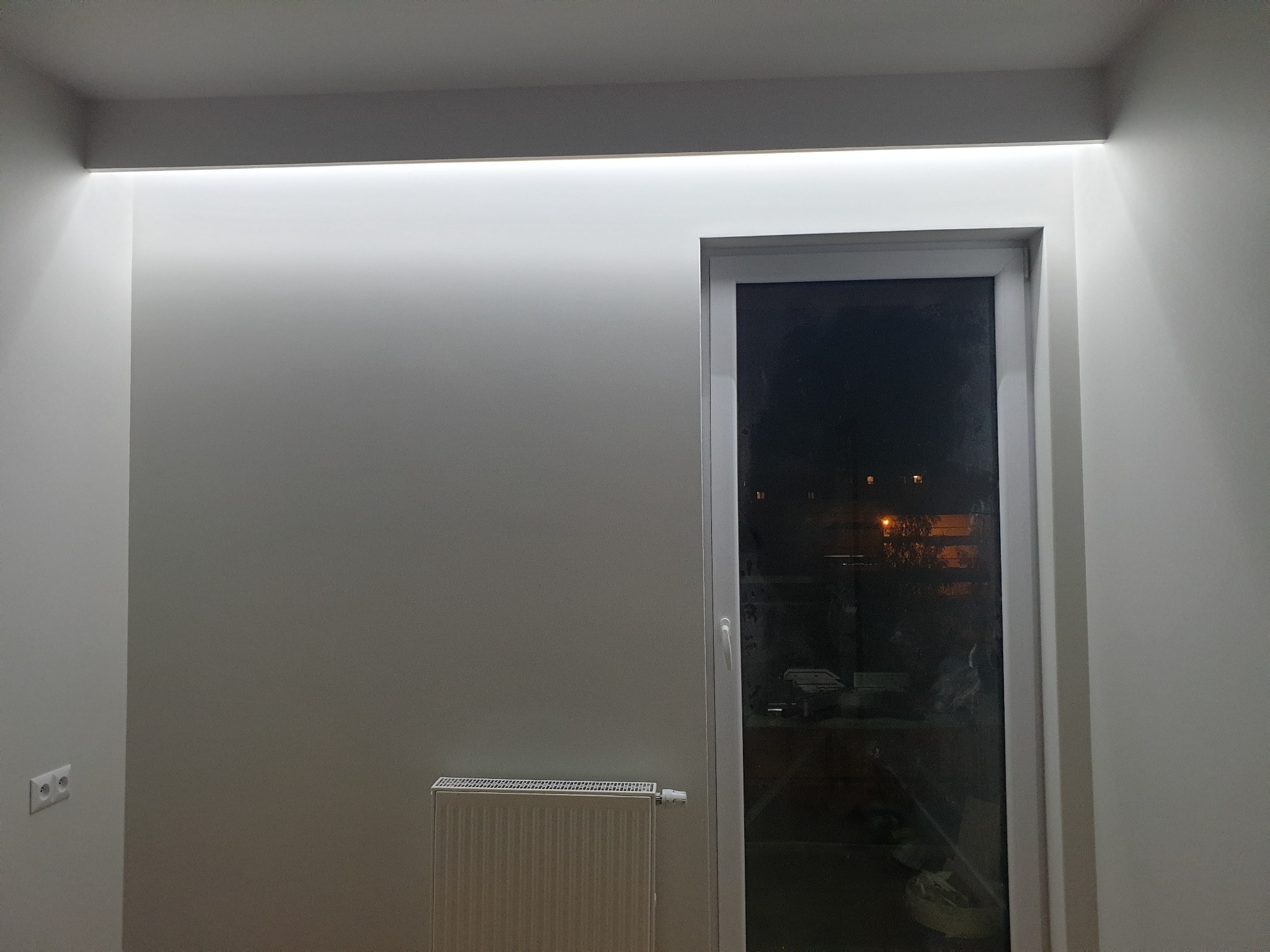 Białe, pomalowane na gładko ściany z oświetleniem LED podsufitowym, okno PCV z widokiem na nocne miasto, grzejnik panelowy pod oknem i gniazdko elektryczne na ścianie.