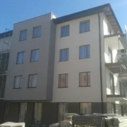 Nowoczesny budynek wielorodzinny po ociepleniu elewacji, z widocznymi rusztowaniami częściowo osłoniętymi siatką ochronną, jasna kolorystyka, błękitne niebo w tle.