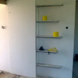 Zabudowa z płyt G-K tworząca półki w trakcie montażu, widoczne narzędzia i materiały budowlane na półkach, surowy stan podłogi.