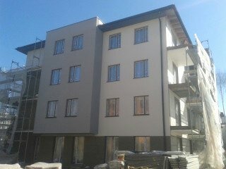 Nowoczesny budynek wielorodzinny po ociepleniu elewacji, z widocznymi rusztowaniami częściowo osłoniętymi siatką ochronną, jasna kolorystyka, błękitne niebo w tle.