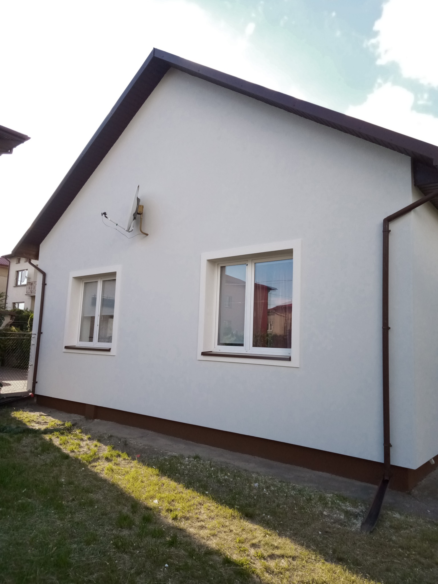 Jasna elewacja domu jednorodzinnego z brązowym wykończeniem dachu i okien, widoczna antena satelitarna, trawnik z cieniami drzew.