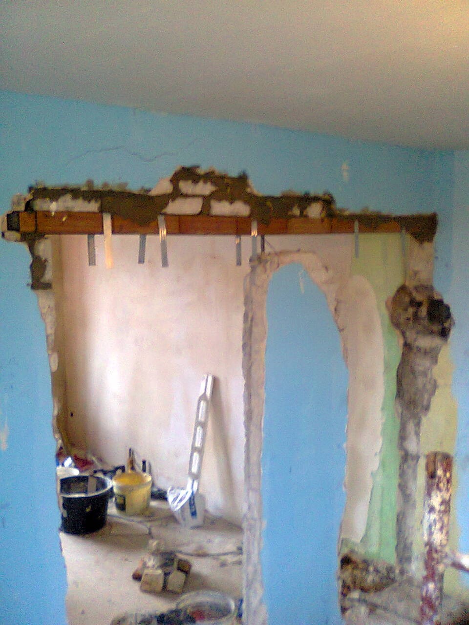 Demontaż ściany działowej z widocznym otworem na drzwi przesuwne, odsłonięta cegła i metalowe elementy mocujące, w tle wiadro i drabina.