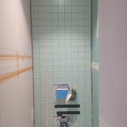 Inglot.glazura kompleksowe usługi glazurnicze - Ściana w trakcie układania jasnoniebieskich płytek w łazience, widoczny laserowy wyznacznik poziomu, elementy instalacji sanitarnej i fragmenty narzędzi.