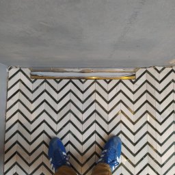 Inglot.glazura kompleksowe usługi glazurnicze - Ukończona podłoga w łazience z białych i zielonych płytek ułożonych we wzór jodełki, widoczne stopy w niebieskich butach sportowych i fragment niedokończonego sufitu podwieszanego.