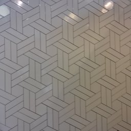 Inglot.glazura kompleksowe usługi glazurnicze - Ściana wyłożona białymi, prostokątnymi płytkami ułożonymi we wzór jodełki, widoczne fugi w kolorze szarym, oświetlenie sufitowe odbija się na powierzchni płytek.