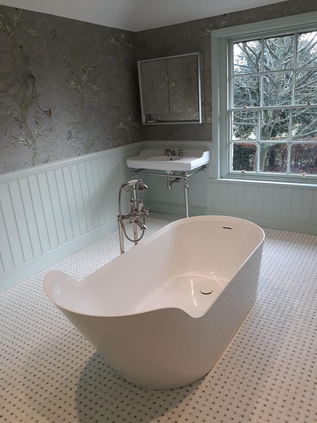 Nowoczesna, wolnostojąca biała wanna w łazience z tapetą w kwiaty i okiennicą w odcieniach szarości, na podłodze jasna mozaika.