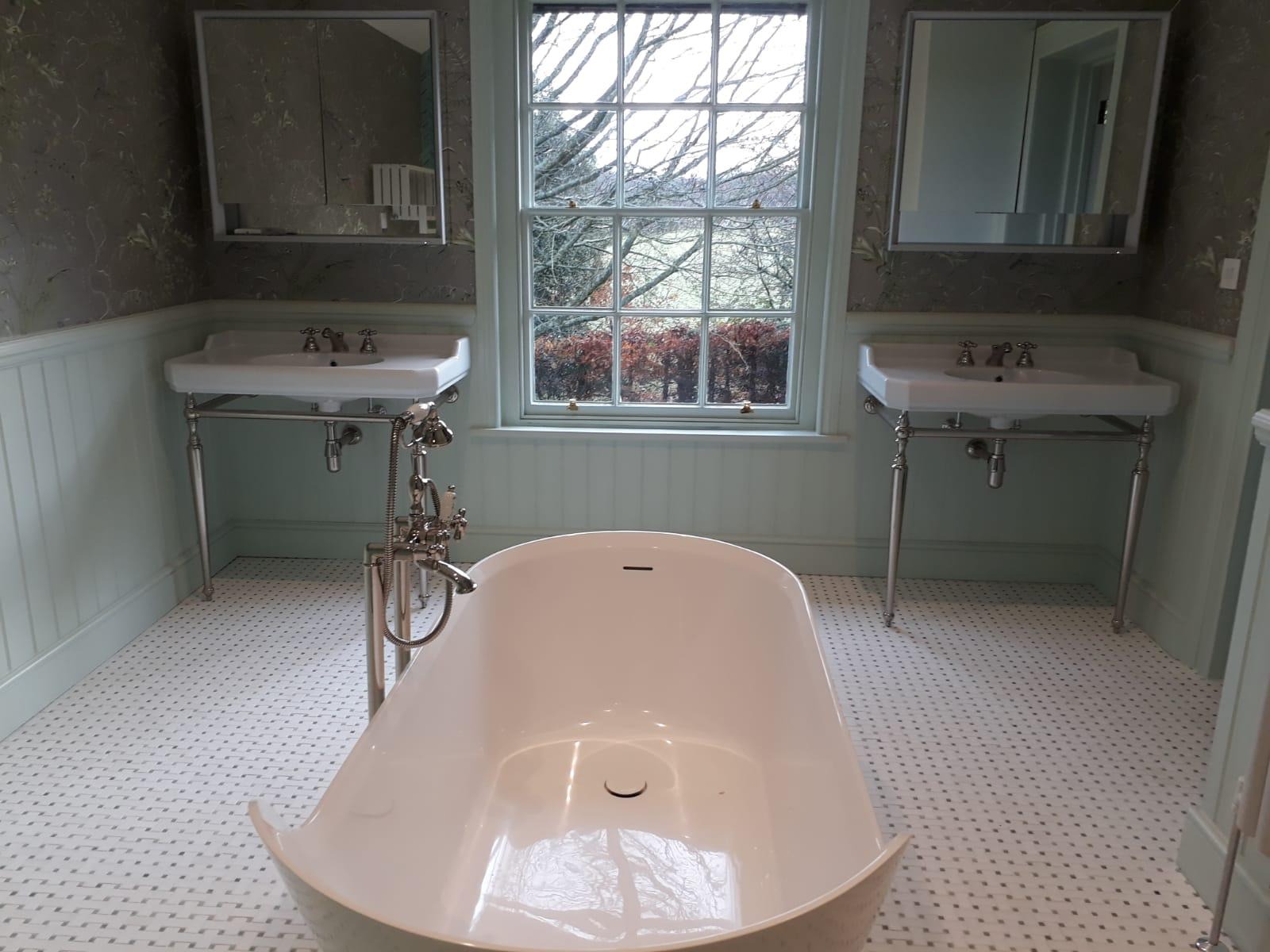 Elegancka łazienka w pastelowych odcieniach z wolnostojącą wanną, dwoma umywalkami na metalowych nogach oraz oknem z widokiem na zewnątrz.