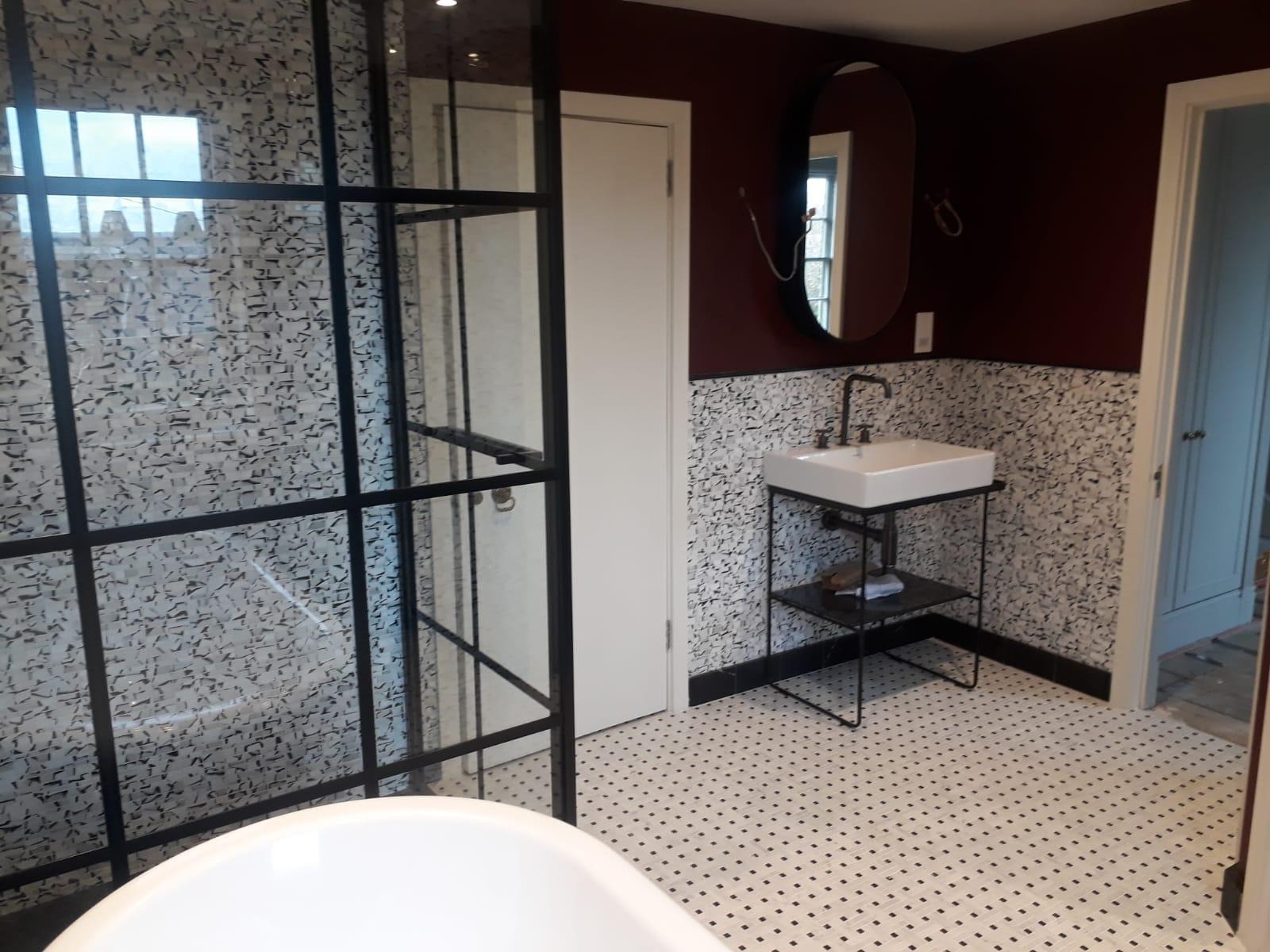 Nowoczesna łazienka z kabiną prysznicową typu walk-in z czarnymi szprosami, białą umywalką na metalowym stelażu, owalnym lustrem i podłogą w biało-czarną mozaikę.