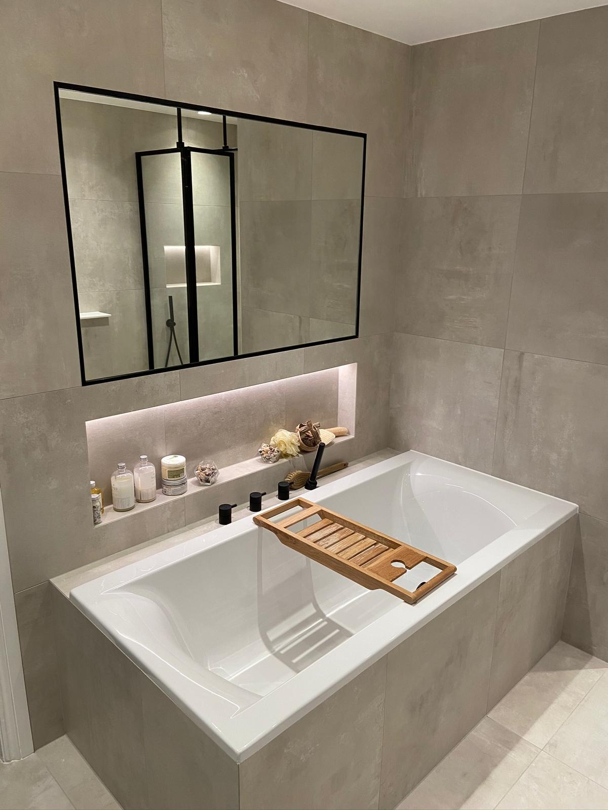 Nowoczesna łazienka z wanną obłożoną płytkami imitującymi beton, drewnianą nakładką na wannę, wnęką z oświetleniem LED i czarną ramą lustra odbijającego kabinę prysznicową.