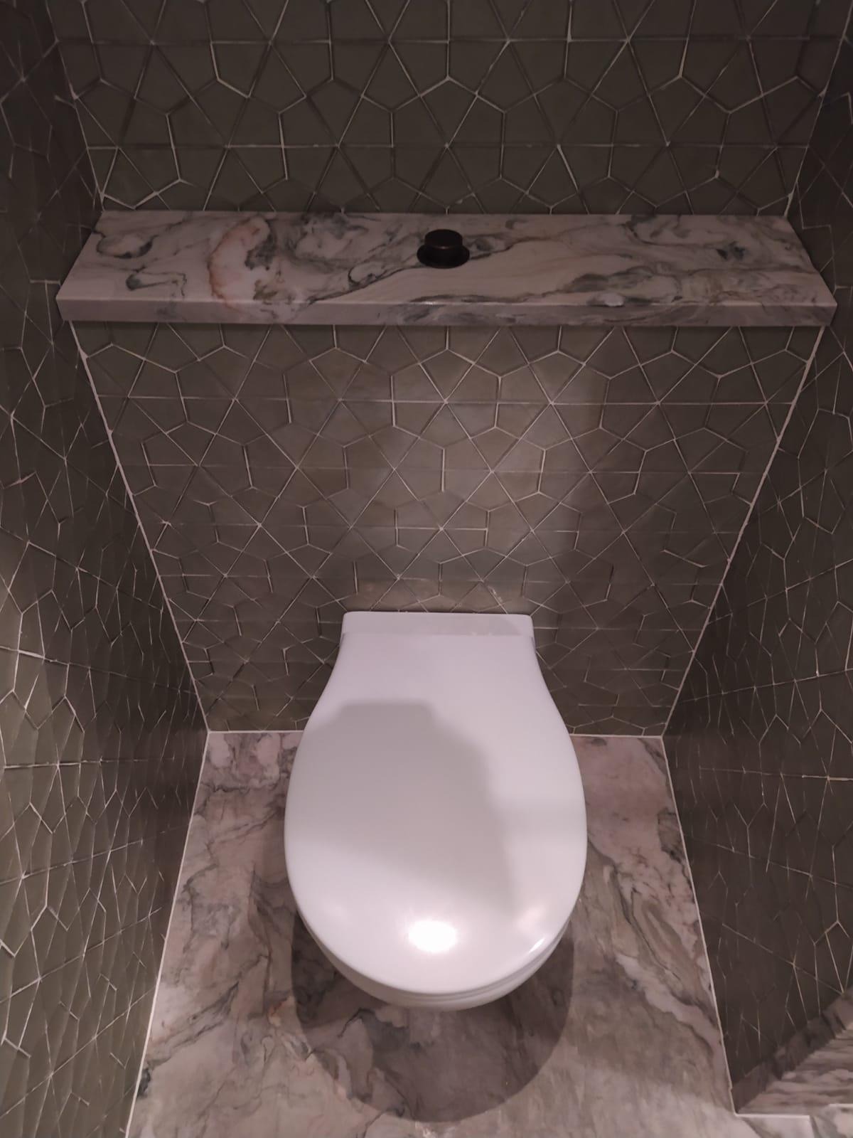 Nowoczesna toaleta w łazience wykończonej szarą mozaiką o geometrycznym wzorze, uzupełniona marmurową półką nad spłuczką i podłogą z tego samego materiału.