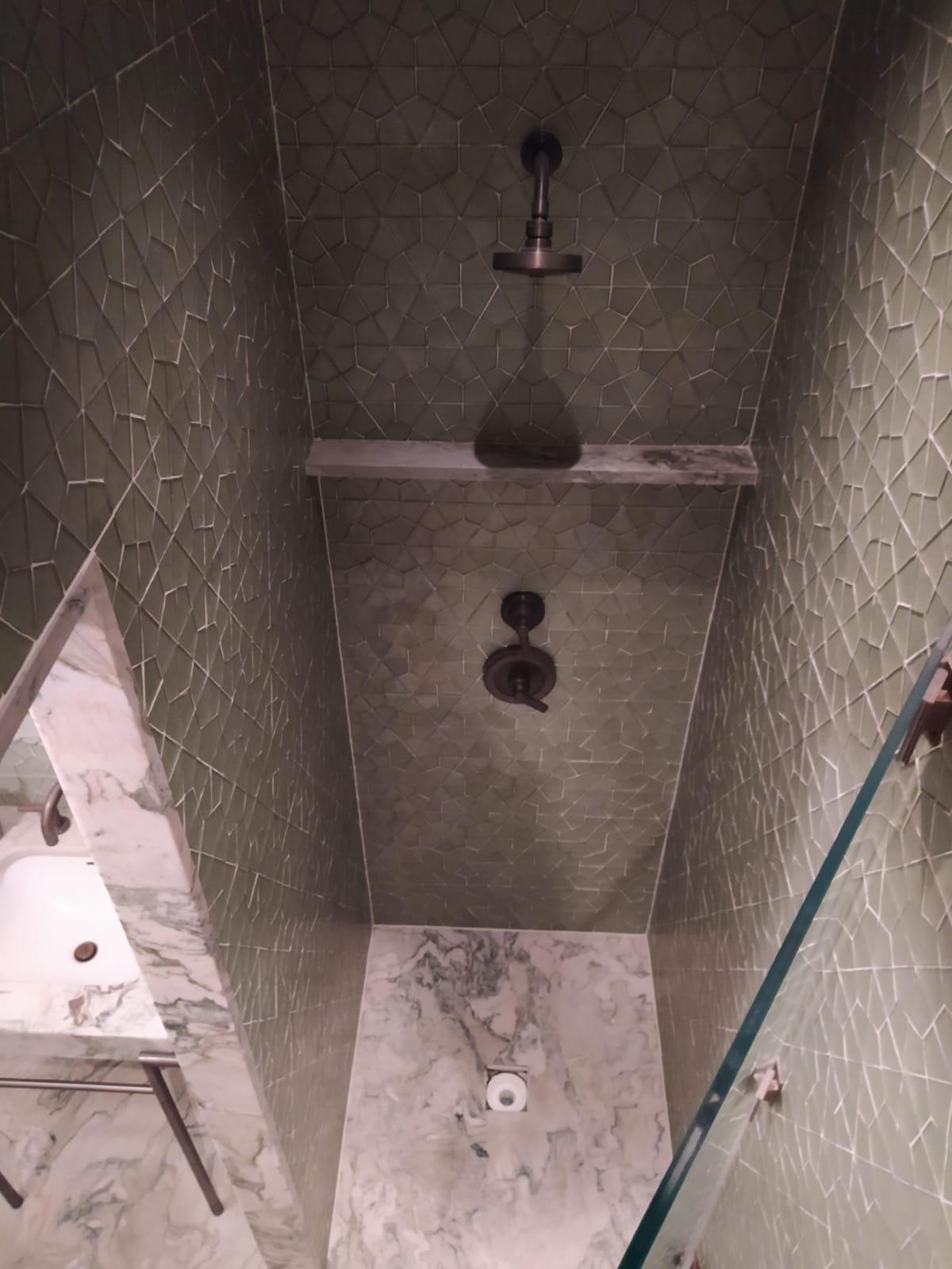 Wnętrze kabiny prysznicowej z zielonymi, geometrycznymi płytkami na ścianach i suficie, widoczne dwie deszczownice w ciemnym kolorze, marmurowa umywalka po lewej stronie i szklana ściana kabiny po...