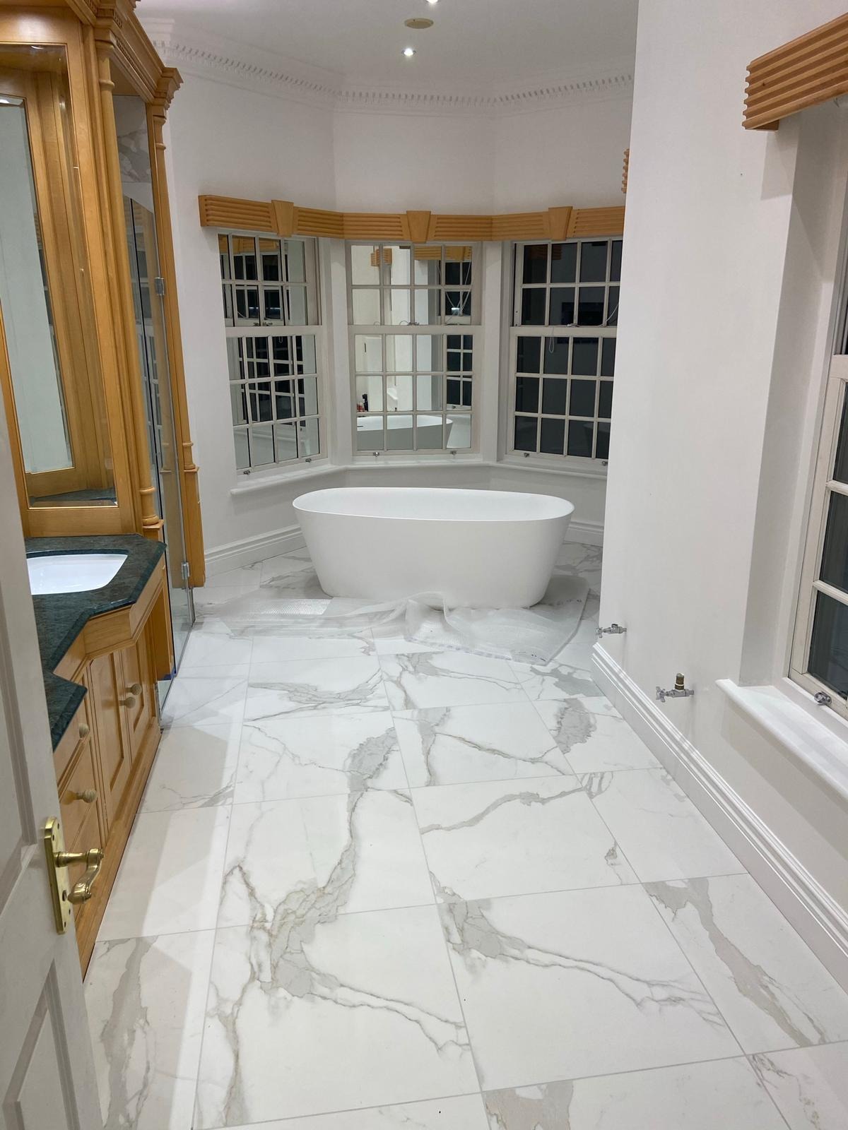 Elegancka łazienka z wolnostojącą wanną na tle okien, marmurowe płytki podłogowe i drewniana szafka z lustrem po lewej stronie.