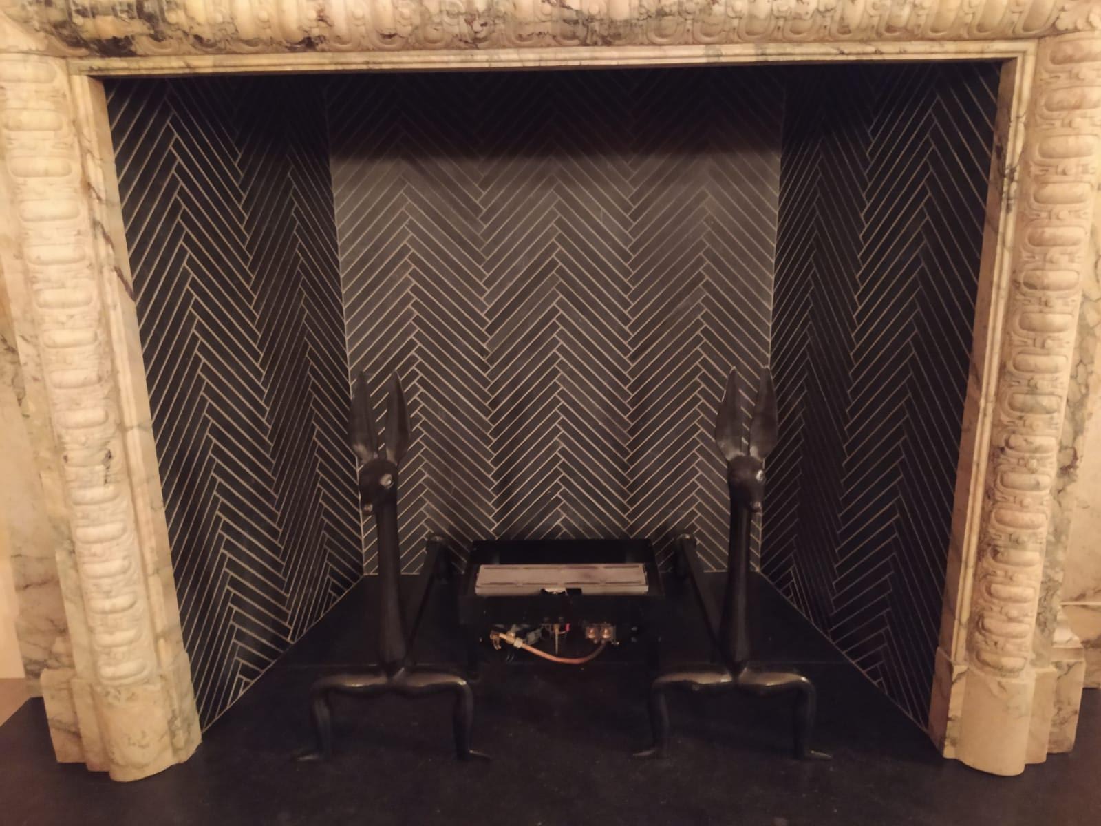 Wykończony kominek z czarną, ułożoną w jodełkę okładziną, flankowany marmurowymi kolumnami i ozdobnymi, metalowymi pogrzebaczami w kształcie głów zwierząt.