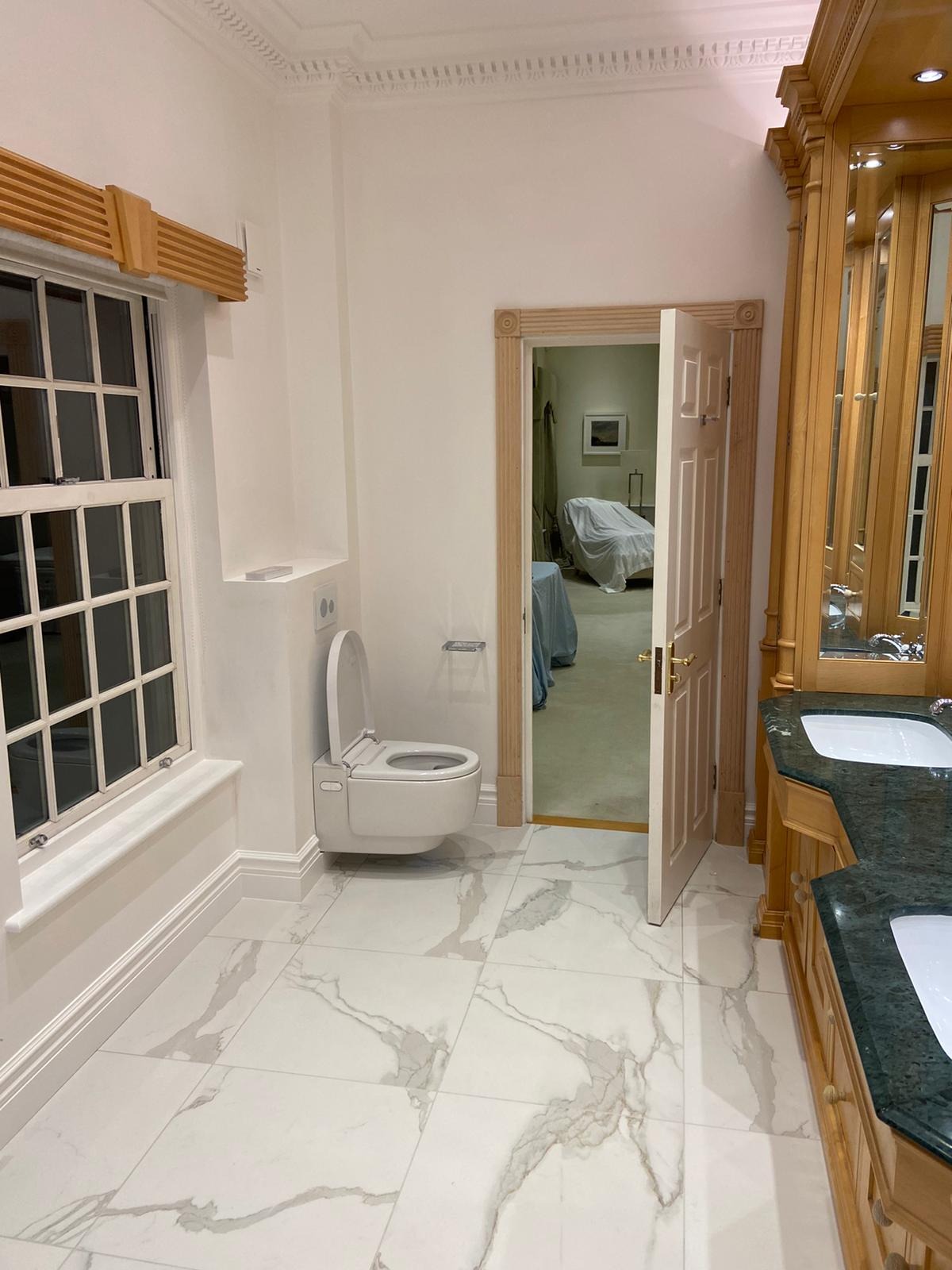 Elegancka, jasna łazienka w Stargardzie z marmurową podłogą, wiszącą toaletą z deską elektroniczną, drewnianą ramą okienną i szafką pod umywalkę z zielonym blatem, widok na otwarte drzwi do pokoju...